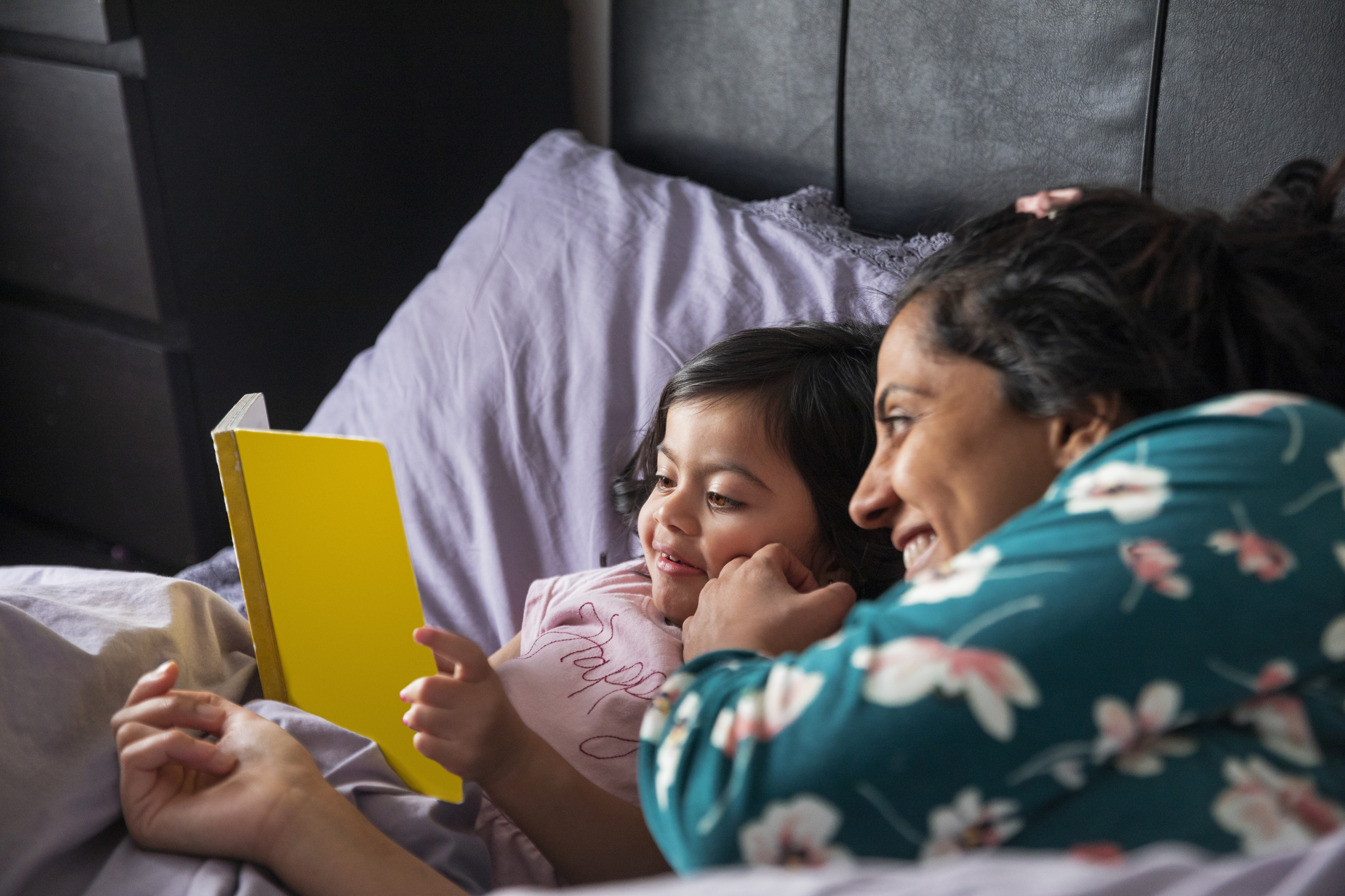 A young girl being read to by her mother in a warm bed.