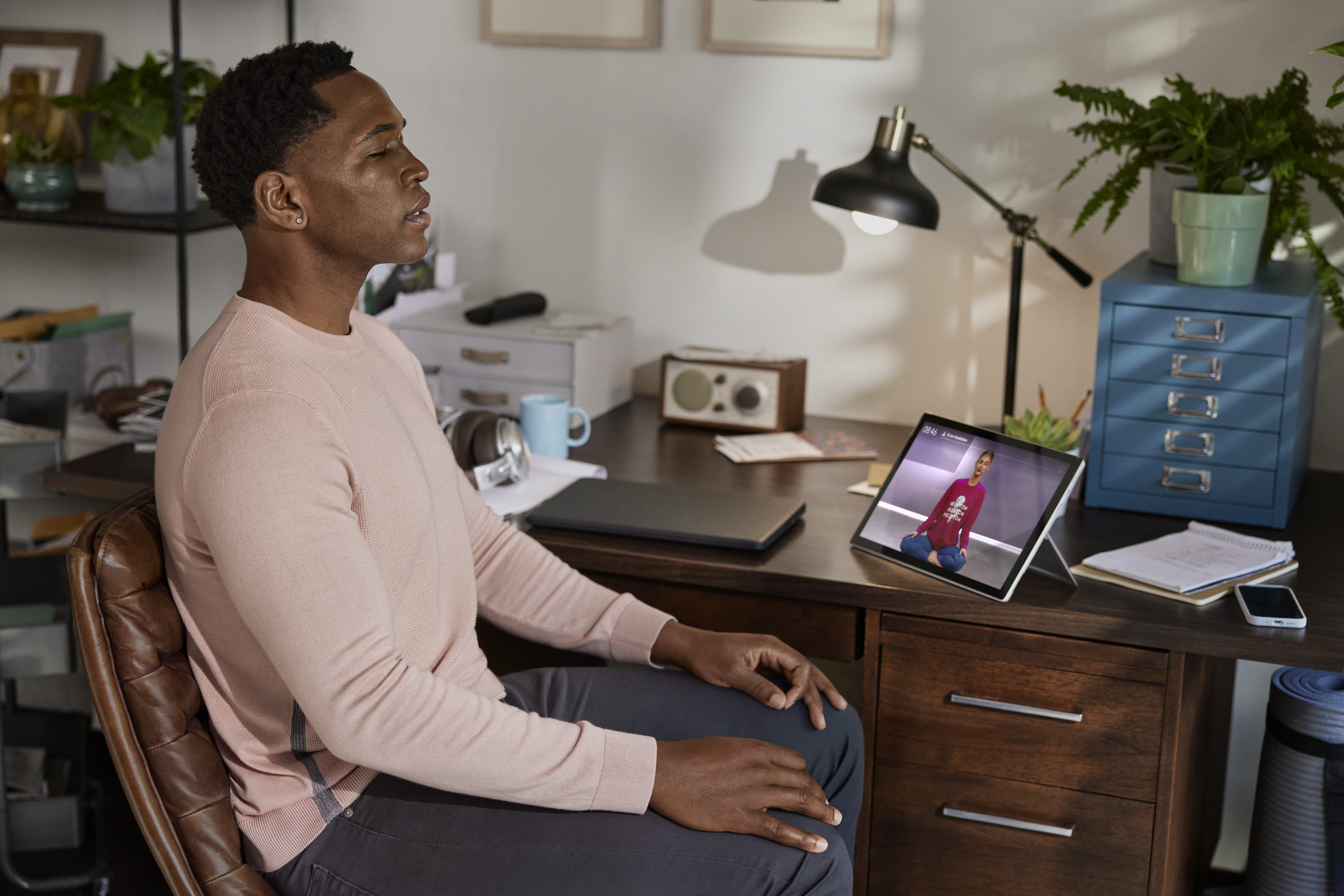 Man, with his eye closed, is performing a Peloton meditation class at his desk at home.