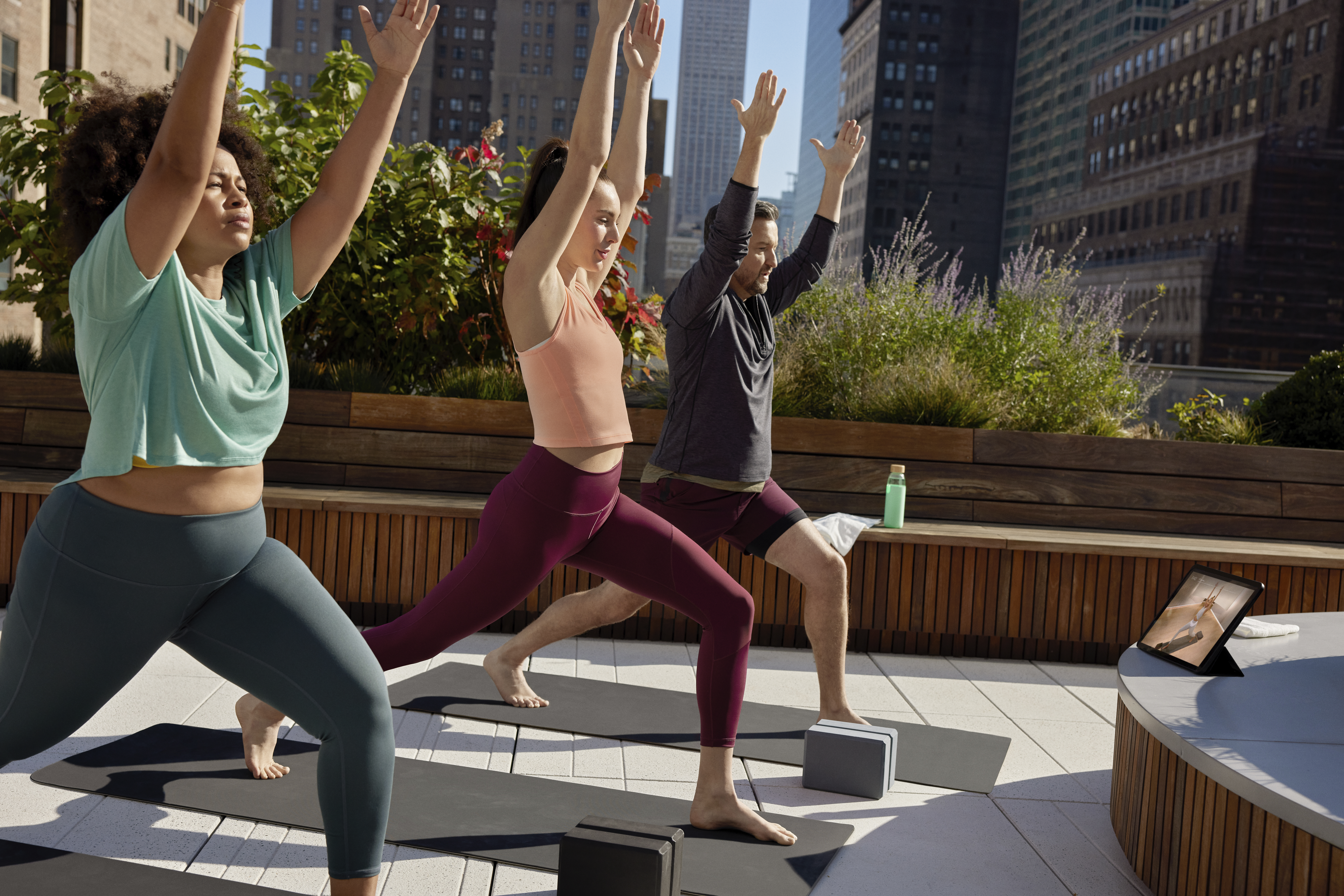 Two women and a man are doing a Peloton yoga class together on the rooftop of their building