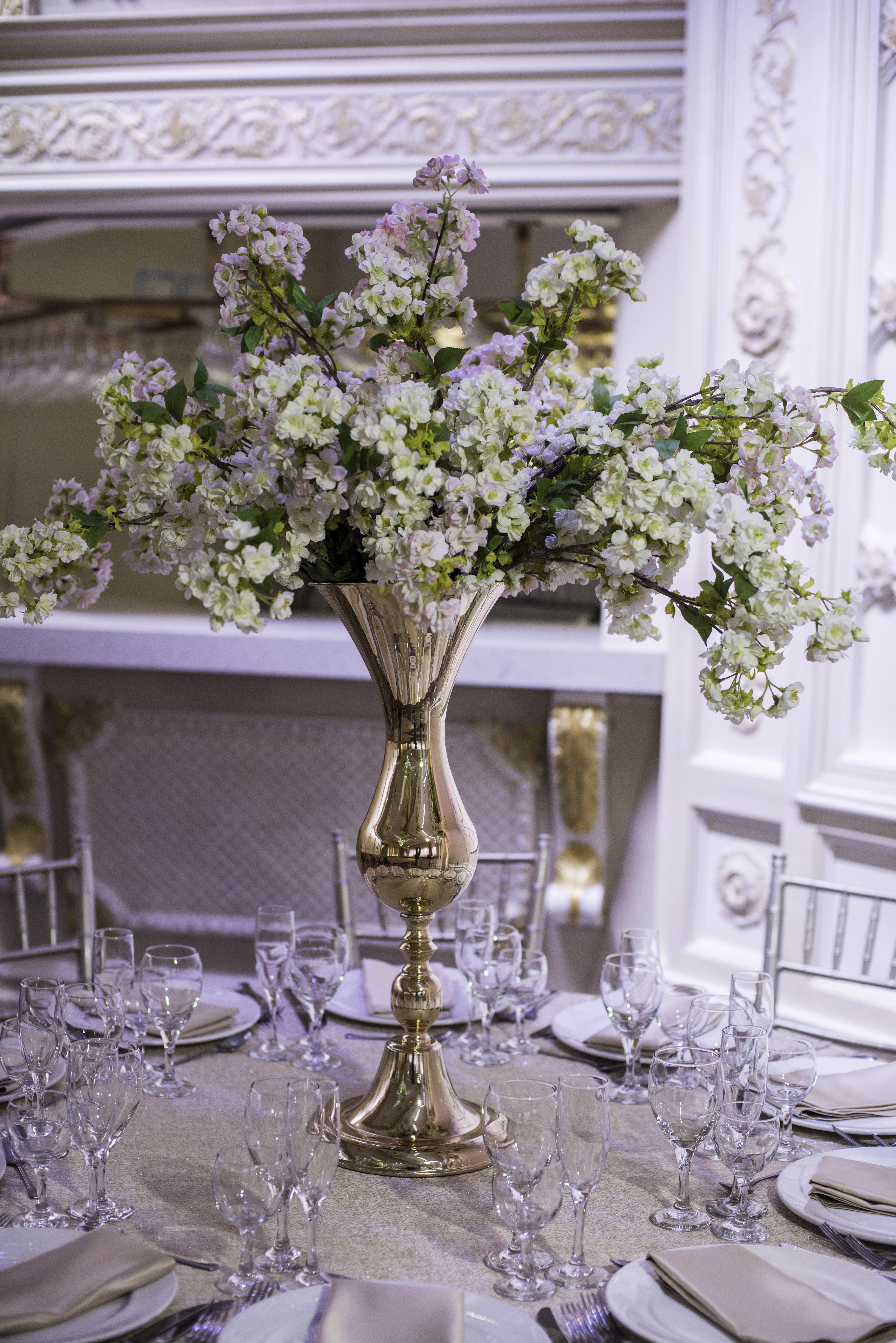 A reflective gold vase with white flowers set on a round table with wedding table setting.