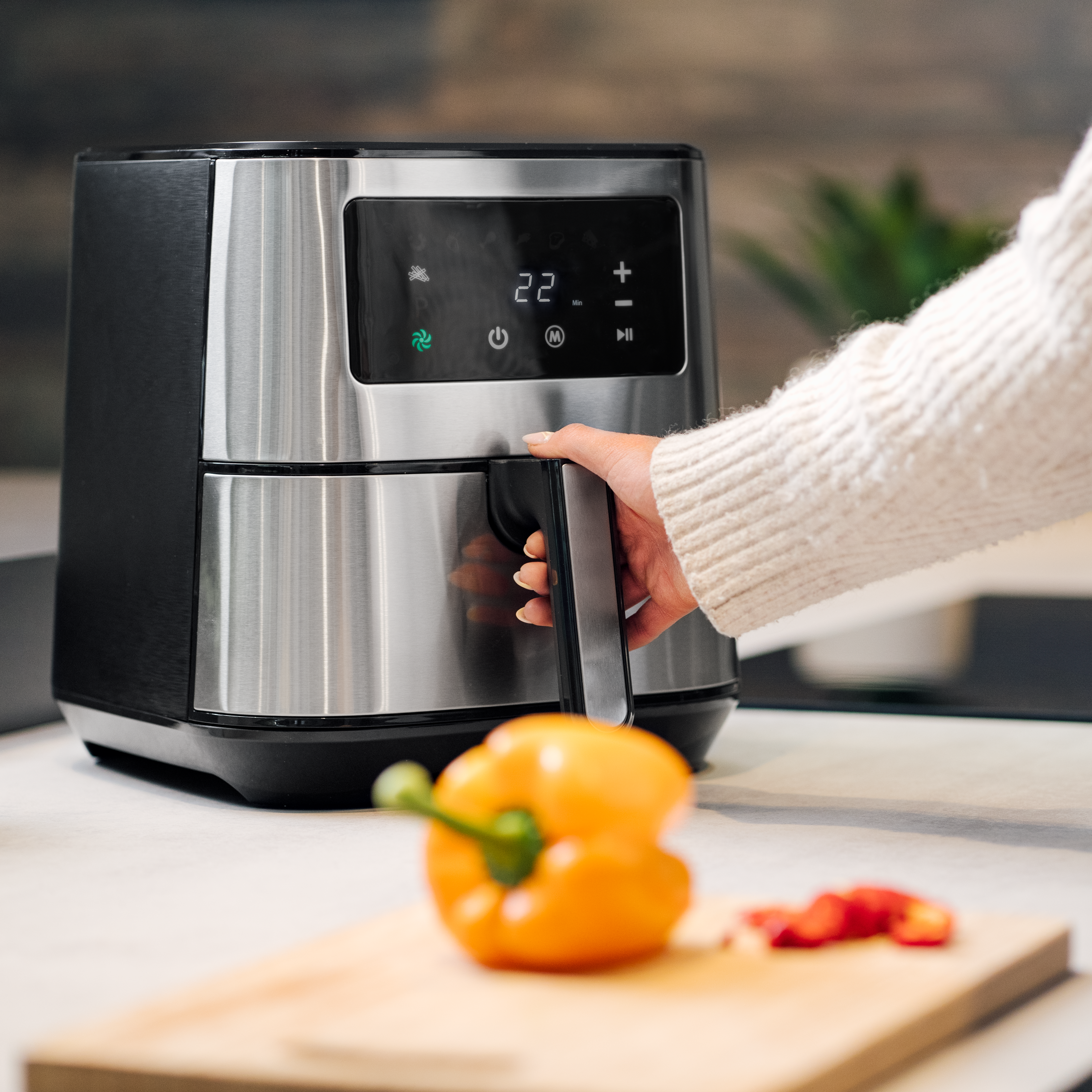 A hand is opening the drawer of a digital air fryer. A yellow bell pepper and sliced red peppers are on a wooden cutting board in the foreground.