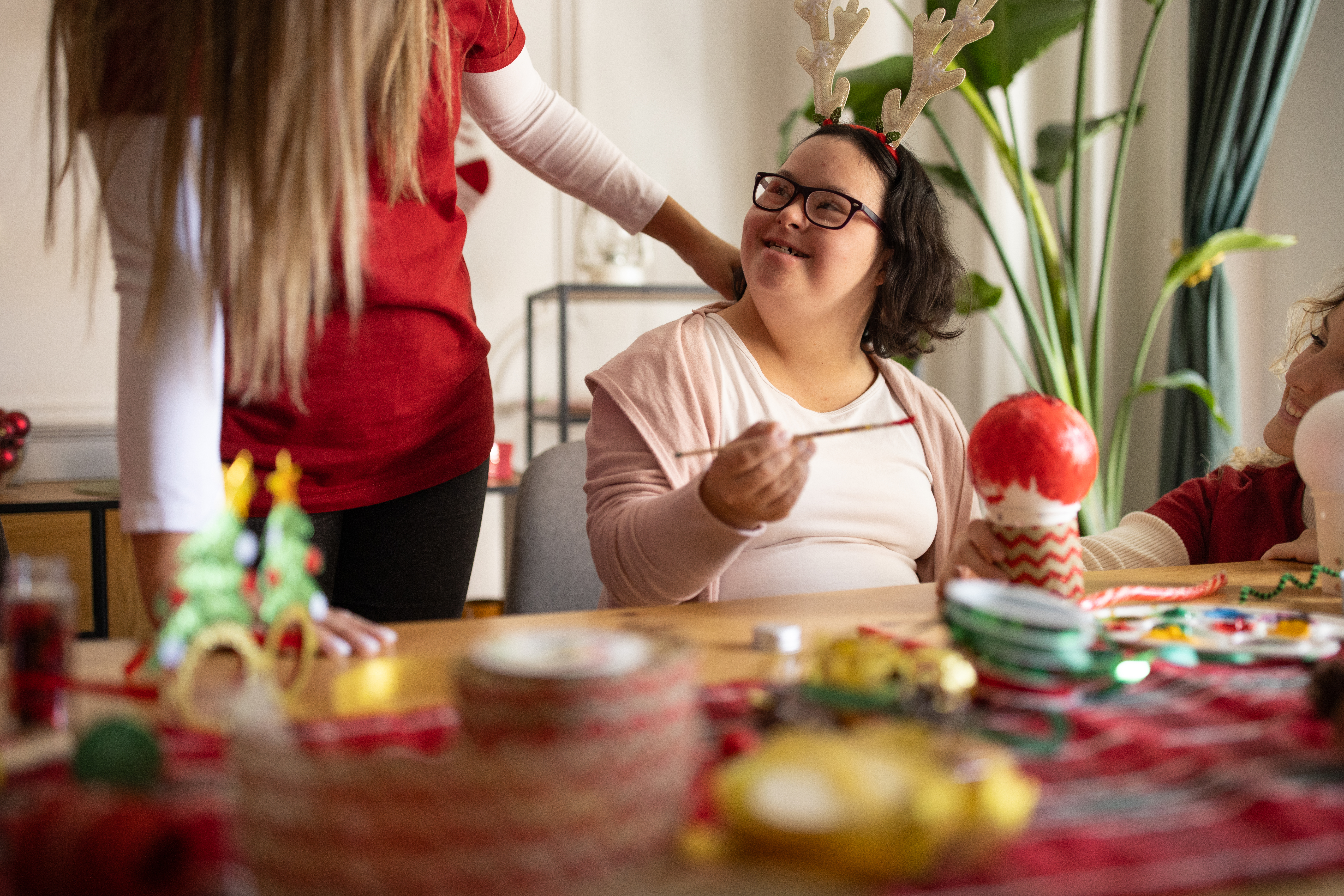 A person seated at a table decorated with festive craft items and ornaments, holding a red object, while another person stands nearby with a hand resting on their shoulder. The table is covered with colorful decorations, ribbons, and holiday-themed items, and there are green plants in the background.