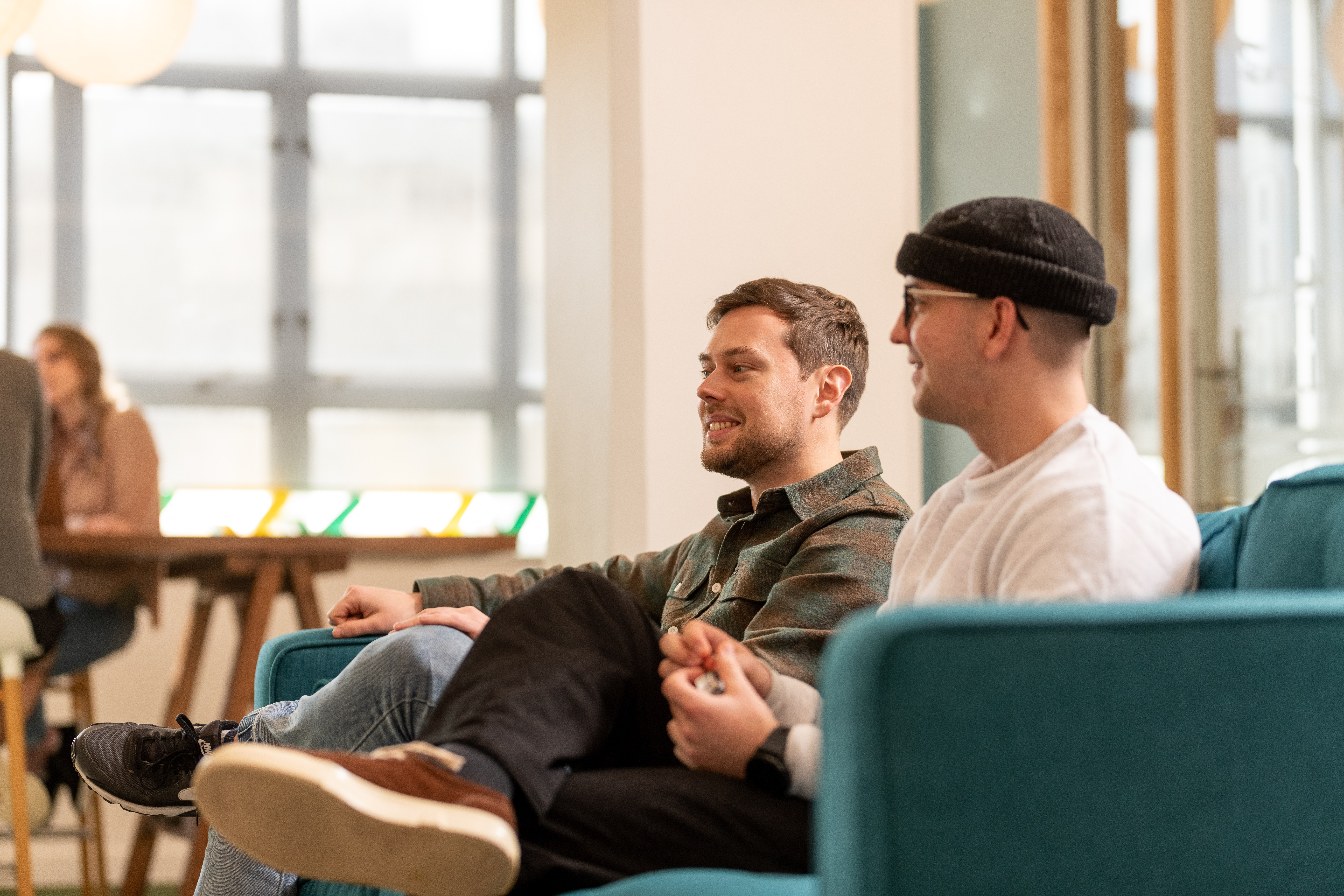 Two men sitting on a sofa. One is wearing a beanie and smiling.