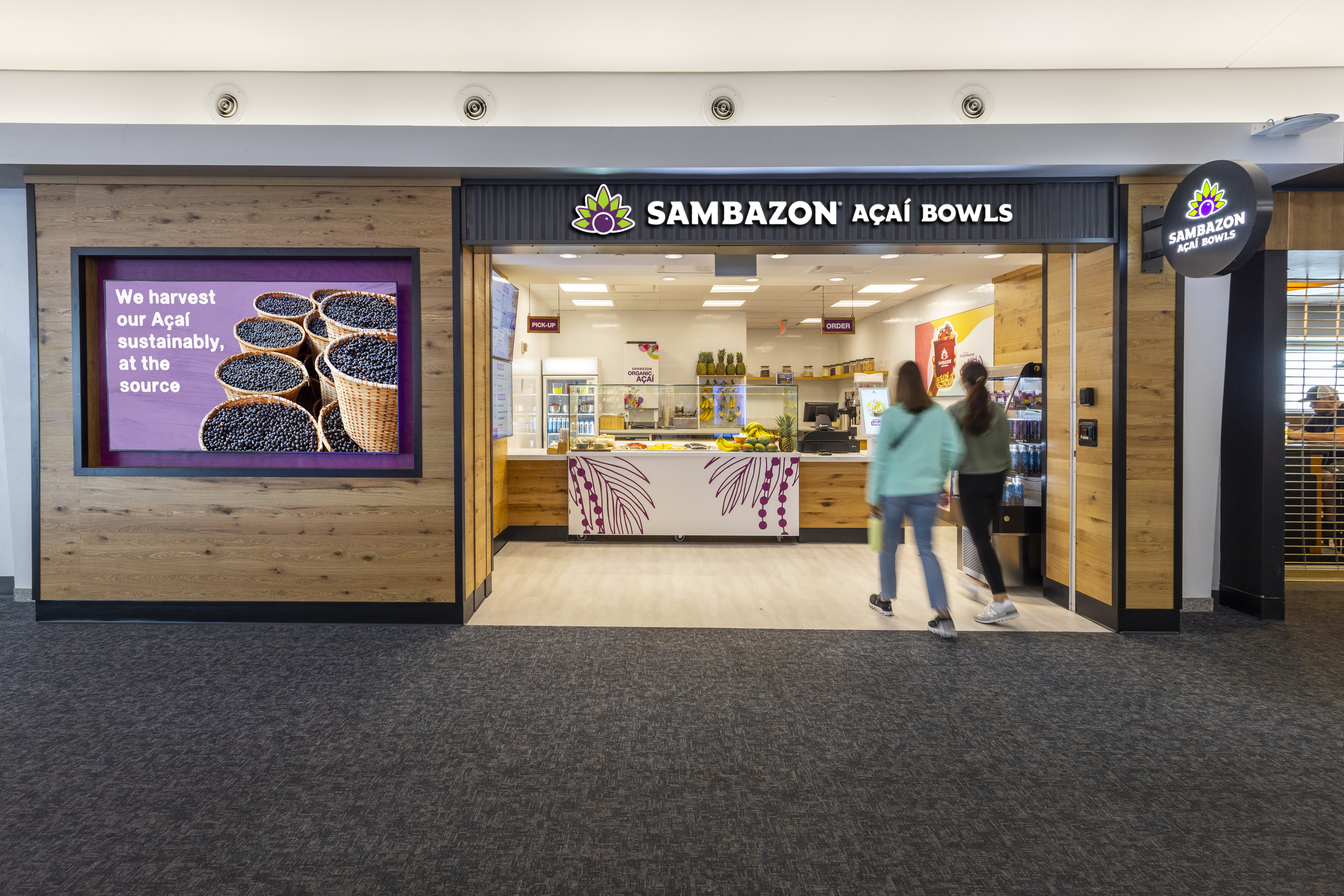 A photo of two people walking into Sambazon at CVG. The storefront is bright and welcoming with wood walls, a counter with an illustration of acai berries in purple down the front. There is an LED screen on the outside left wall of the restaurant.