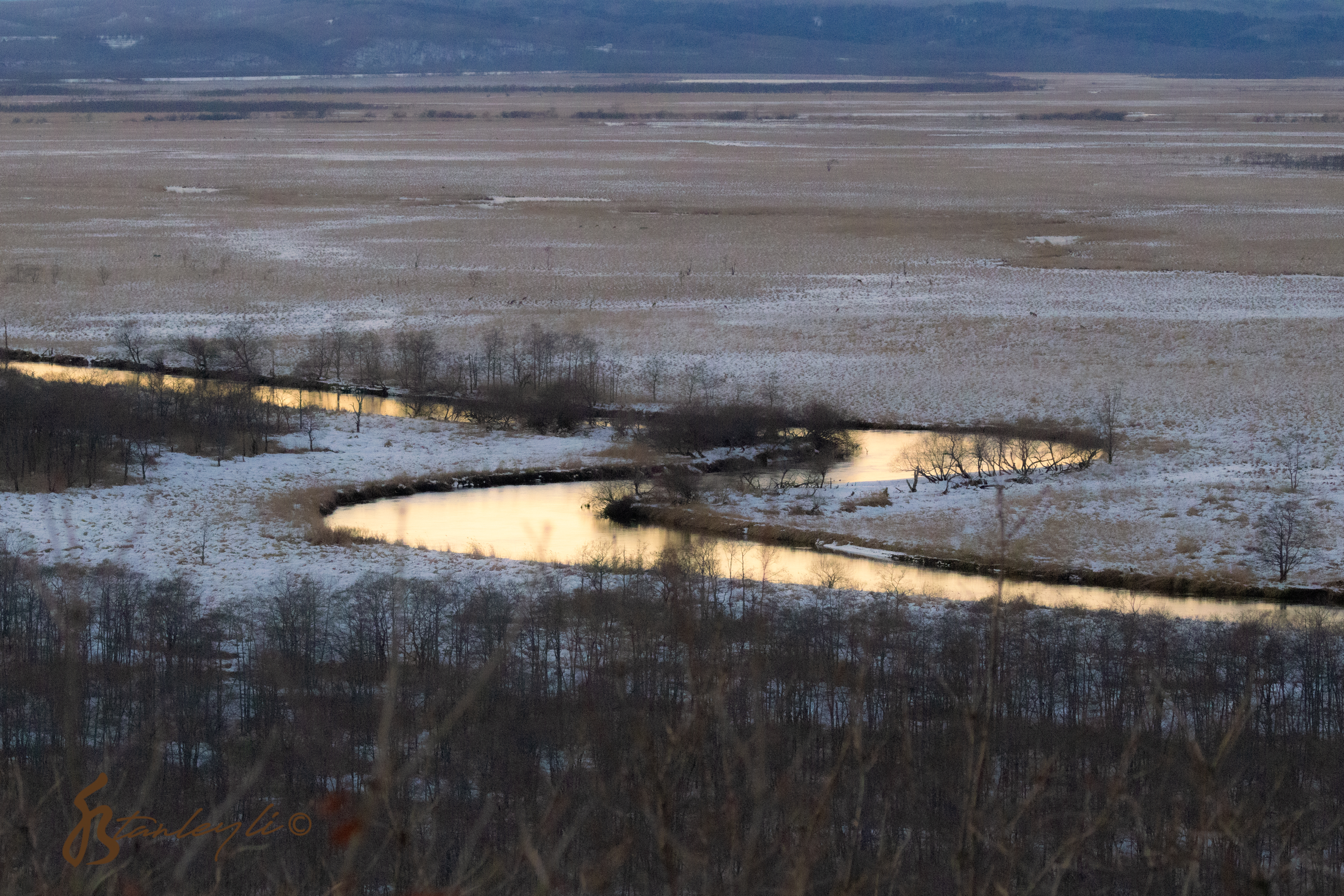 The colours of sunset are reflected in the meandering Kushiro River. The surrounding wetlands is dusted in snow.