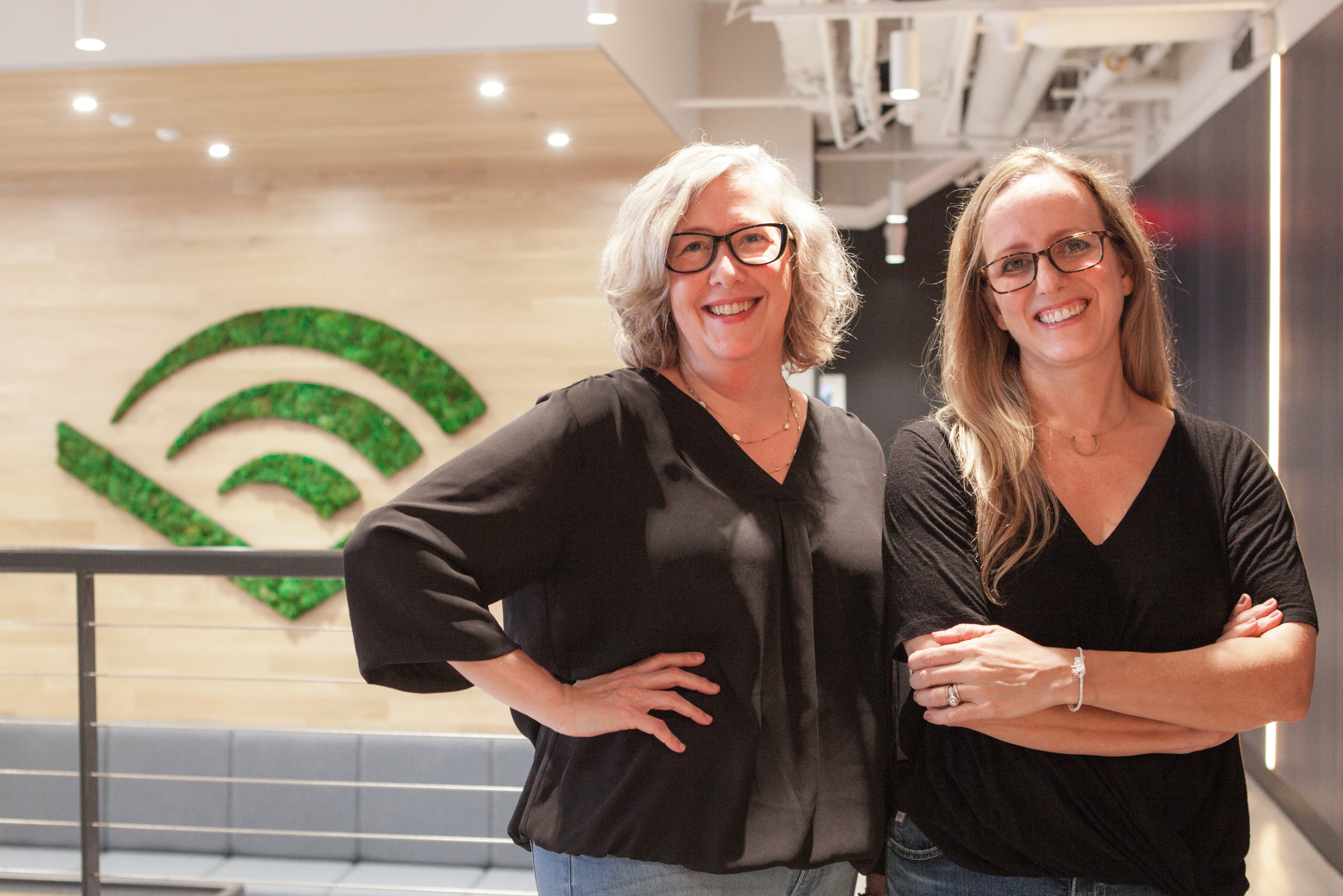 Audible editors Tricia Ford and Emily Cox stand side by side in Audible's office. They are looking directly at the camera and smiling. Both are wearing glasses, black v-neck shirts and jeans. In the background is a staircase and on the wall the Audible chevron carved into the wall in green moss.