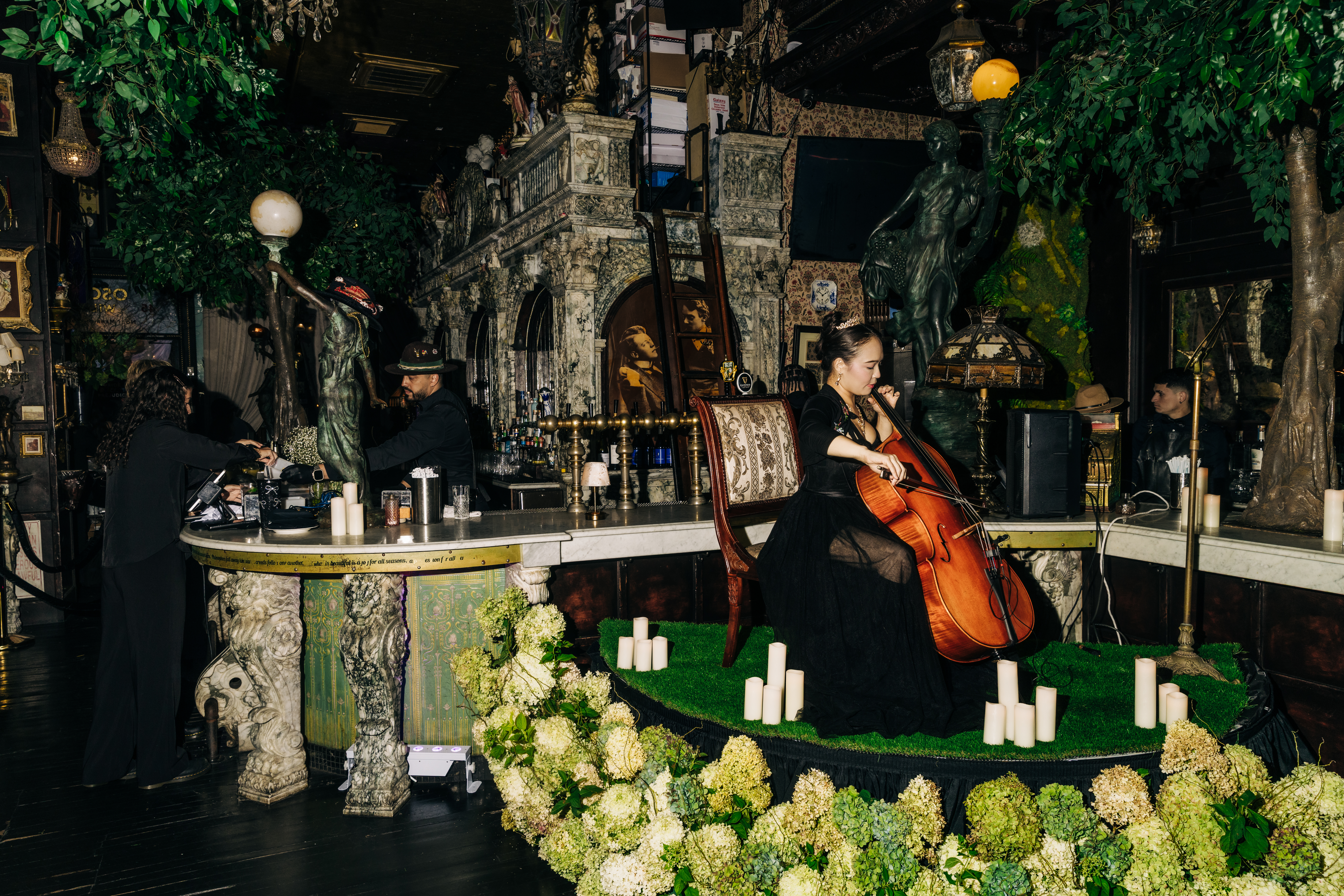 A photo of a cellist playing in a dark, romantically lit room, surrounded by green and white flowers. 