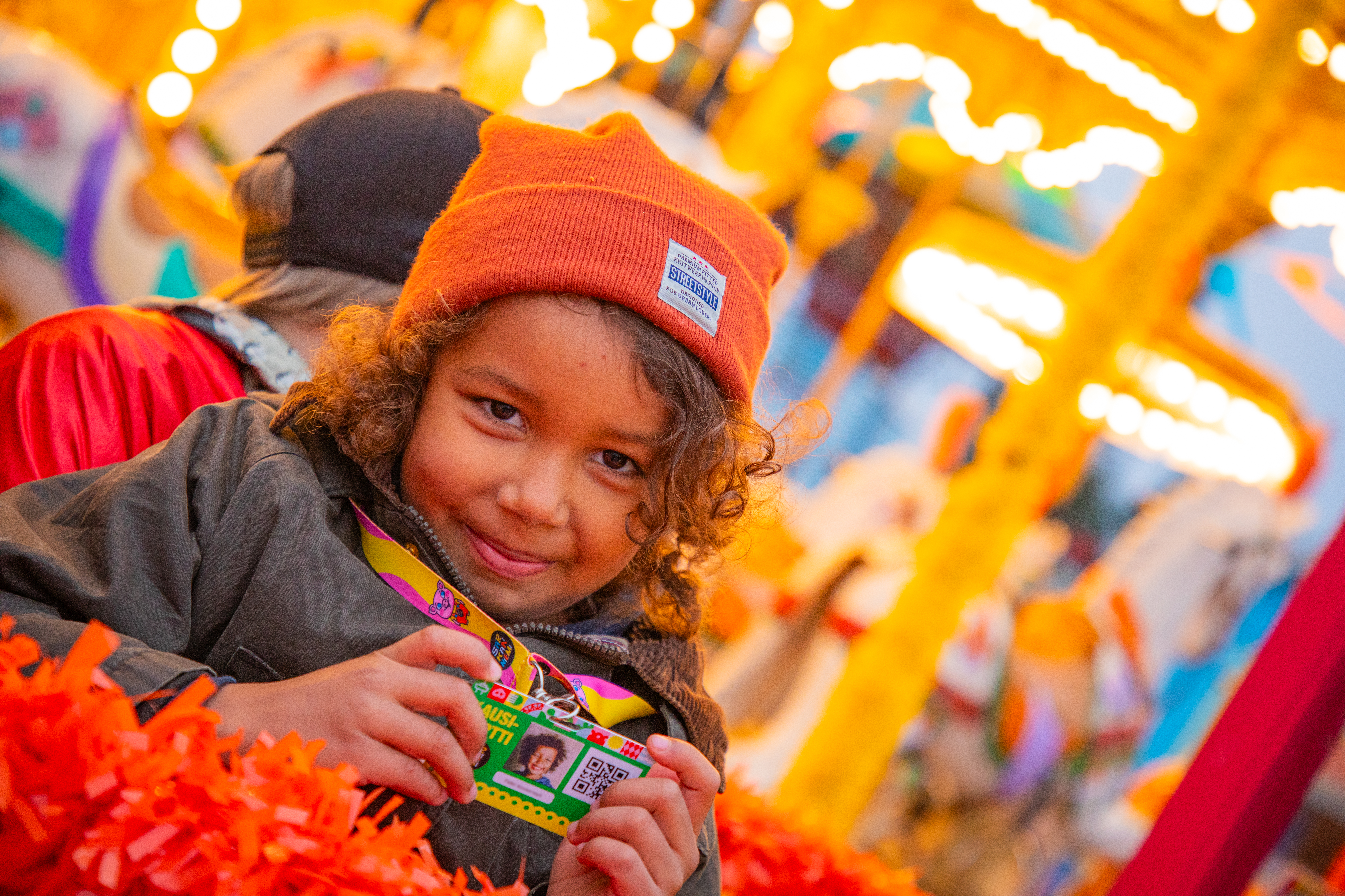 A brown, curly-haired child with an orange hat on their head happily holds the 2024 Season Pass in front of the Särkänniemi Candy Carousel in warm lighting.