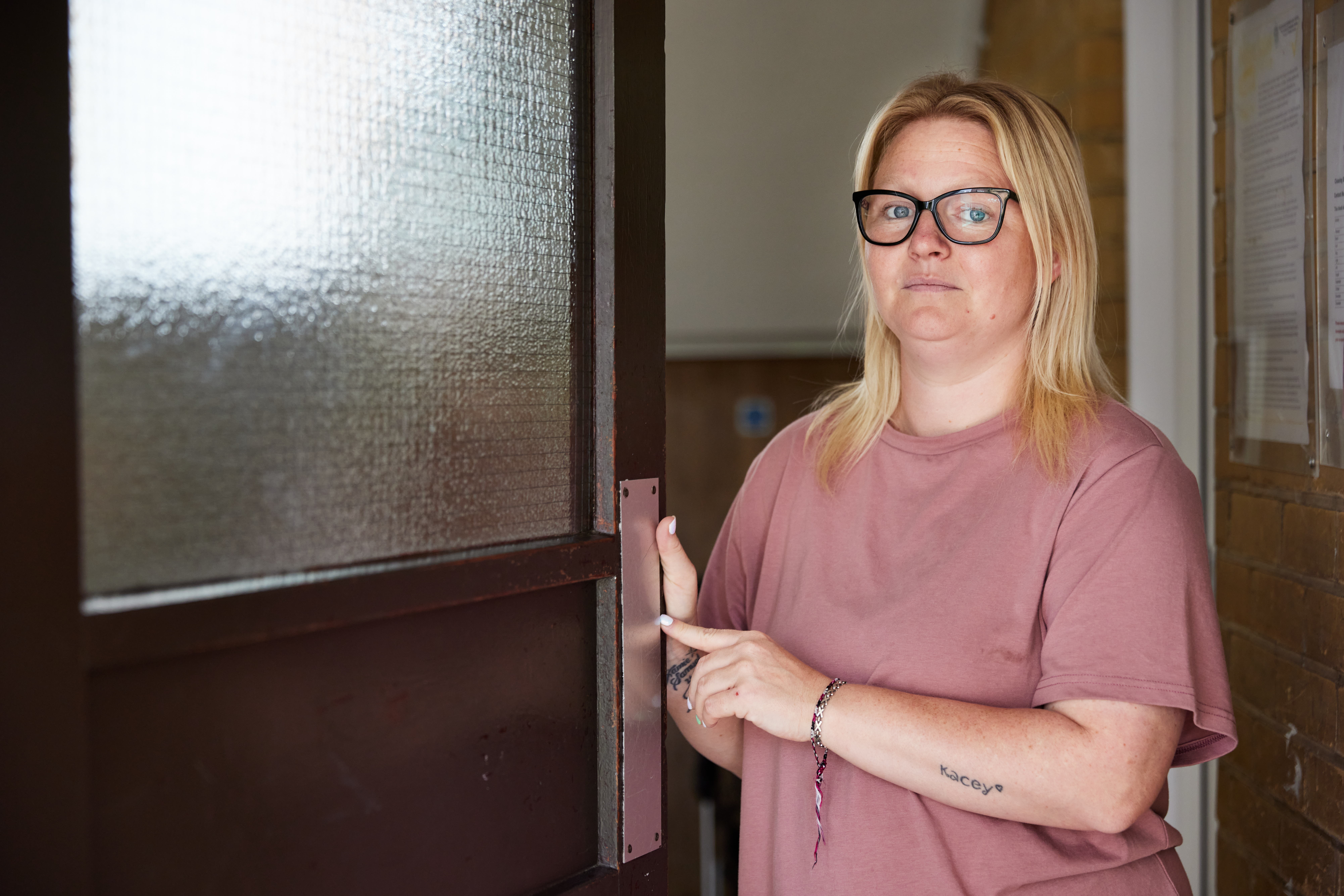 A woman with shoulder-length blonde hair and glasses wearing a pink t-shirt stands at an open door.
