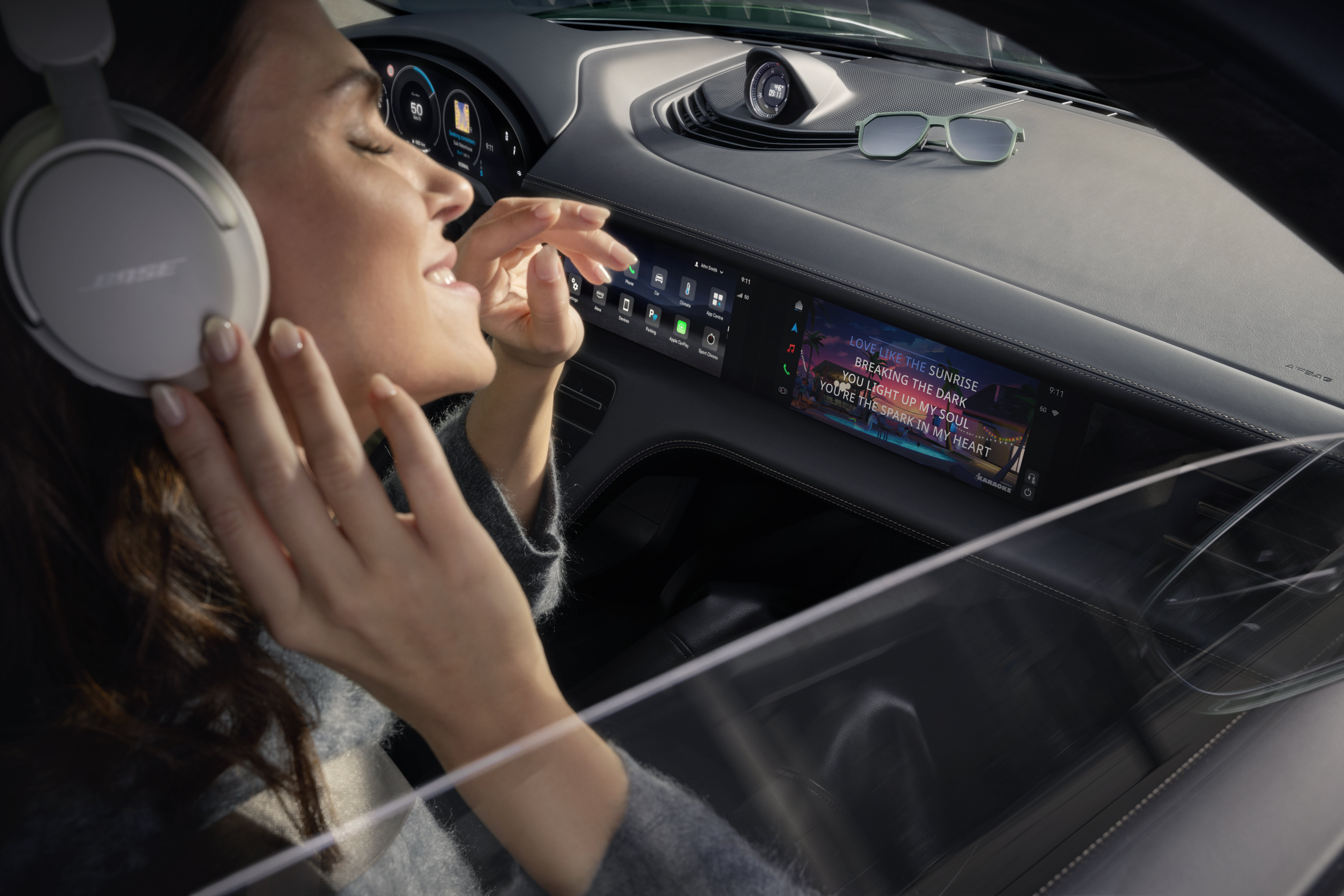 A woman wearing Bose headphones enjoys music inside a Porsche, smiling with her eyes closed and hand raised to her ear. The dashboard display shows karaoke-style lyrics on the passenger screen, with lines like “LOVE LIKE THE SUNRISE” and “YOU LIGHT UP MY SOUL.” Sunglasses rest on the dashboard, and the car's digital interface is visible in the background, along with a speedometer reading 50 mph.