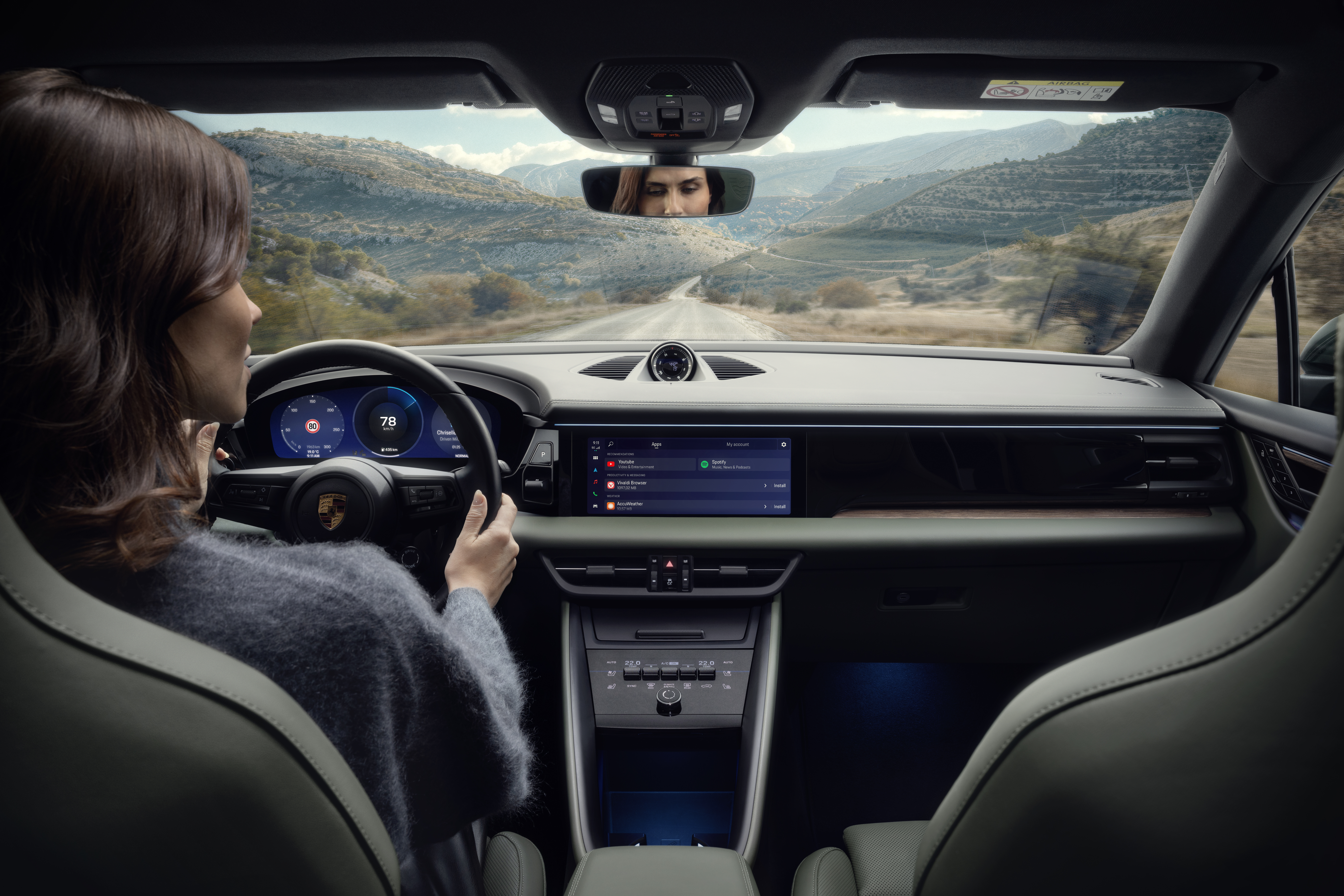 A woman is driving a modern car with a digital cockpit and central touchscreen display. She looks focused on the road ahead, which winds through a scenic mountain landscape. Her face is visible in the rearview mirror, while the dashboard shows the Porsche logo, navigation data, and a music app.