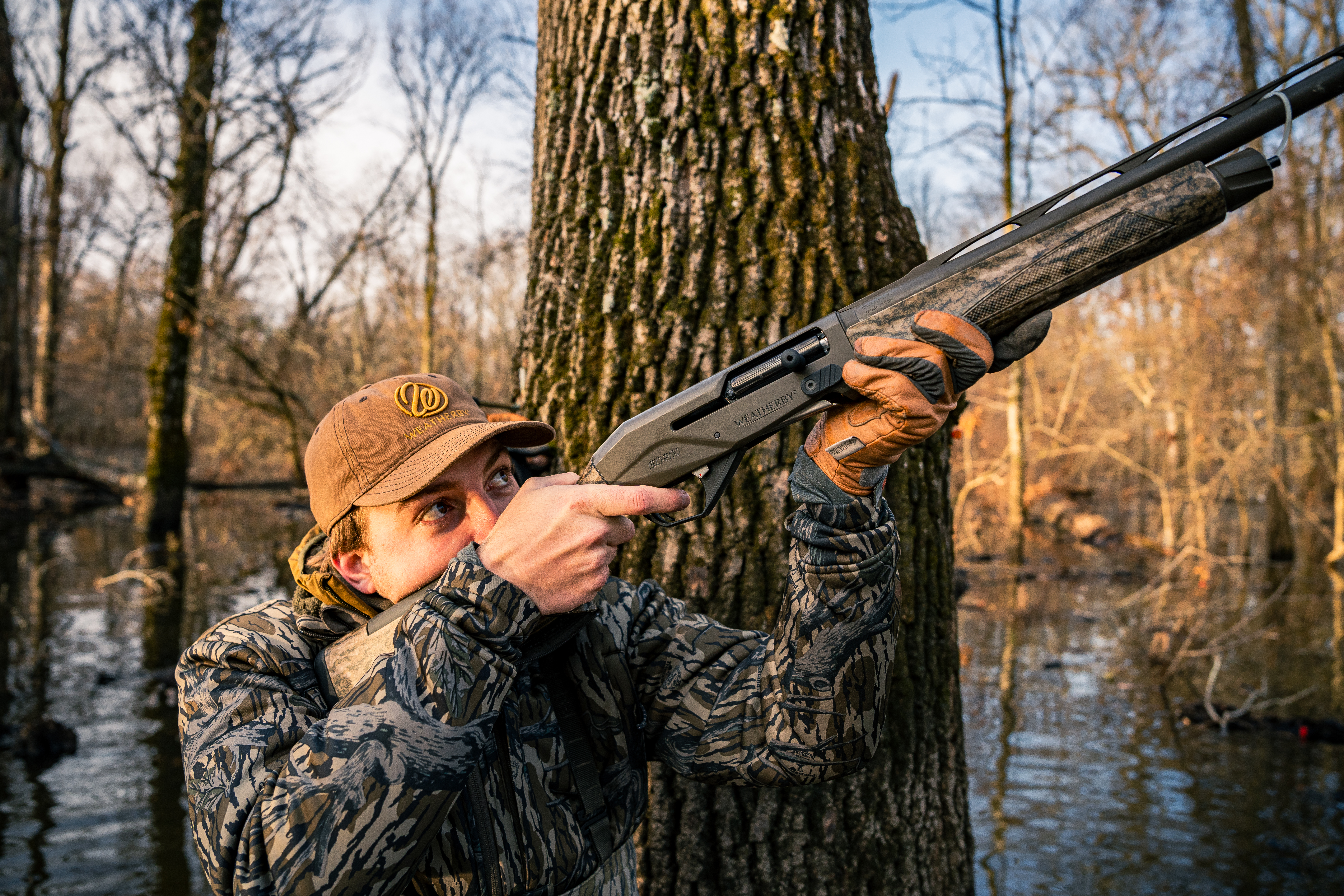 A hunter standing in flooded timber pointing a shotgun towards the sky