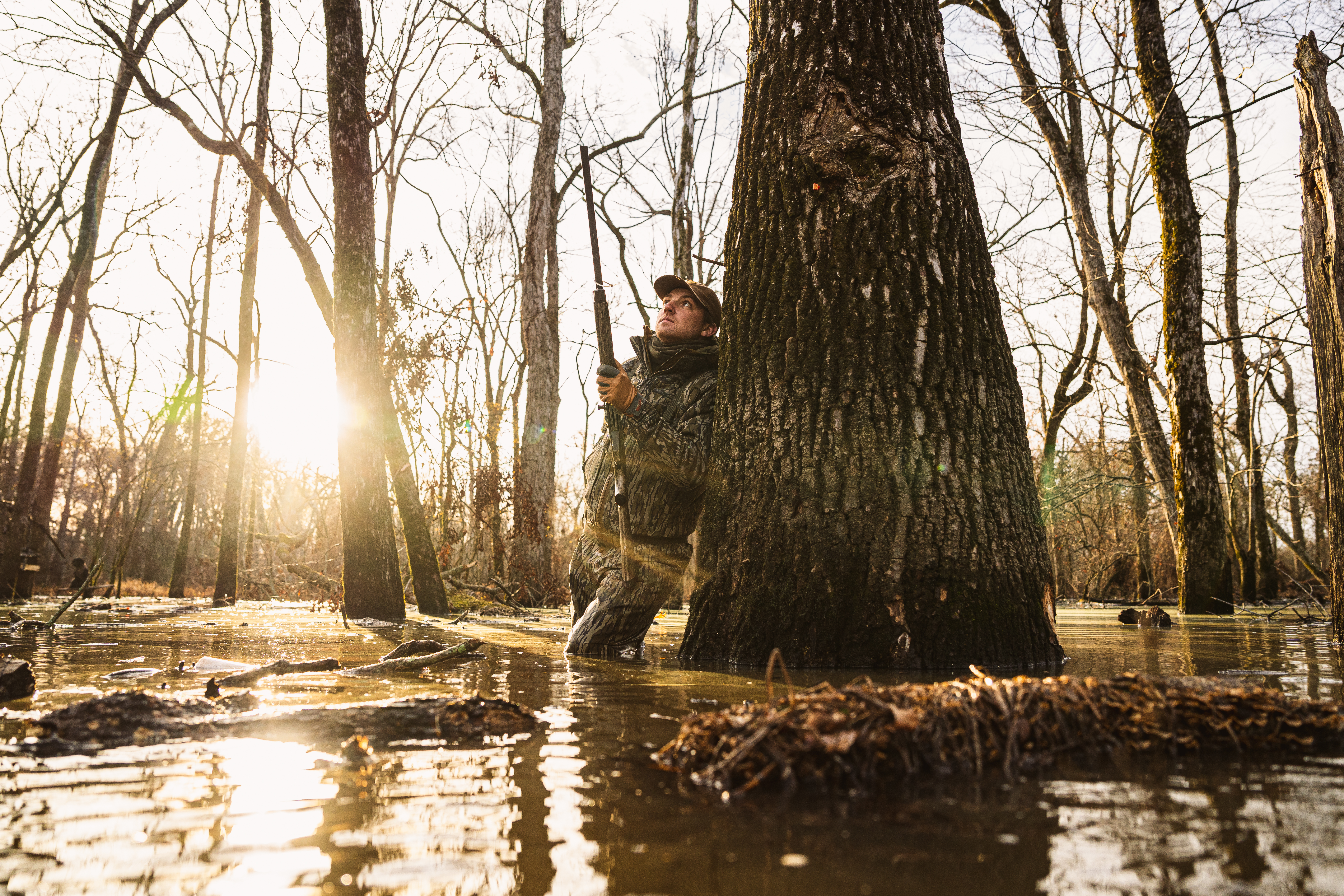 A duck hunter stands in flooded timber leaning up against a tree