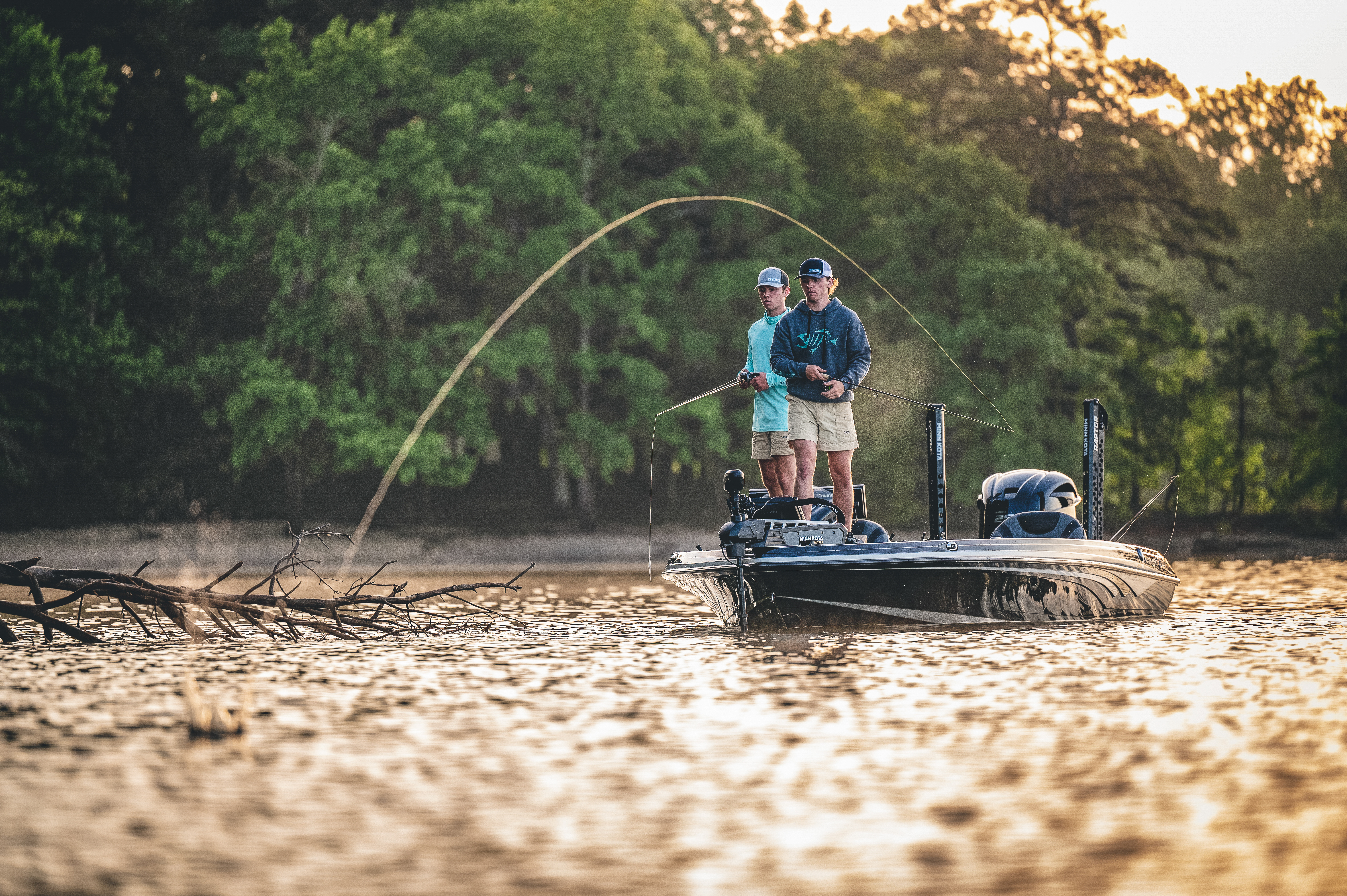 Two bass anglers in a boat cast to a shoreline on a lake.