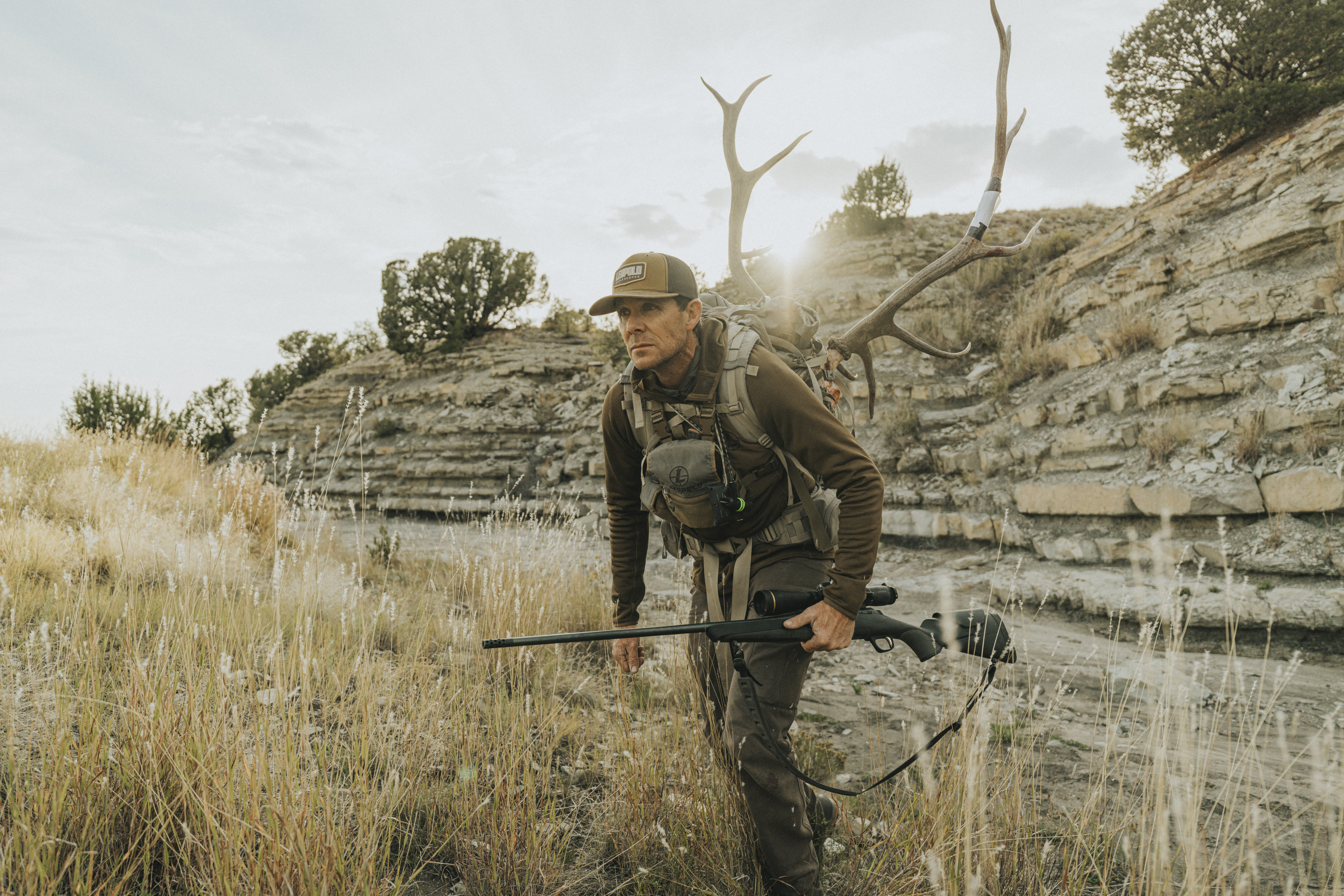 Hunter packs elk antlers out of the field with rifle in hand.  