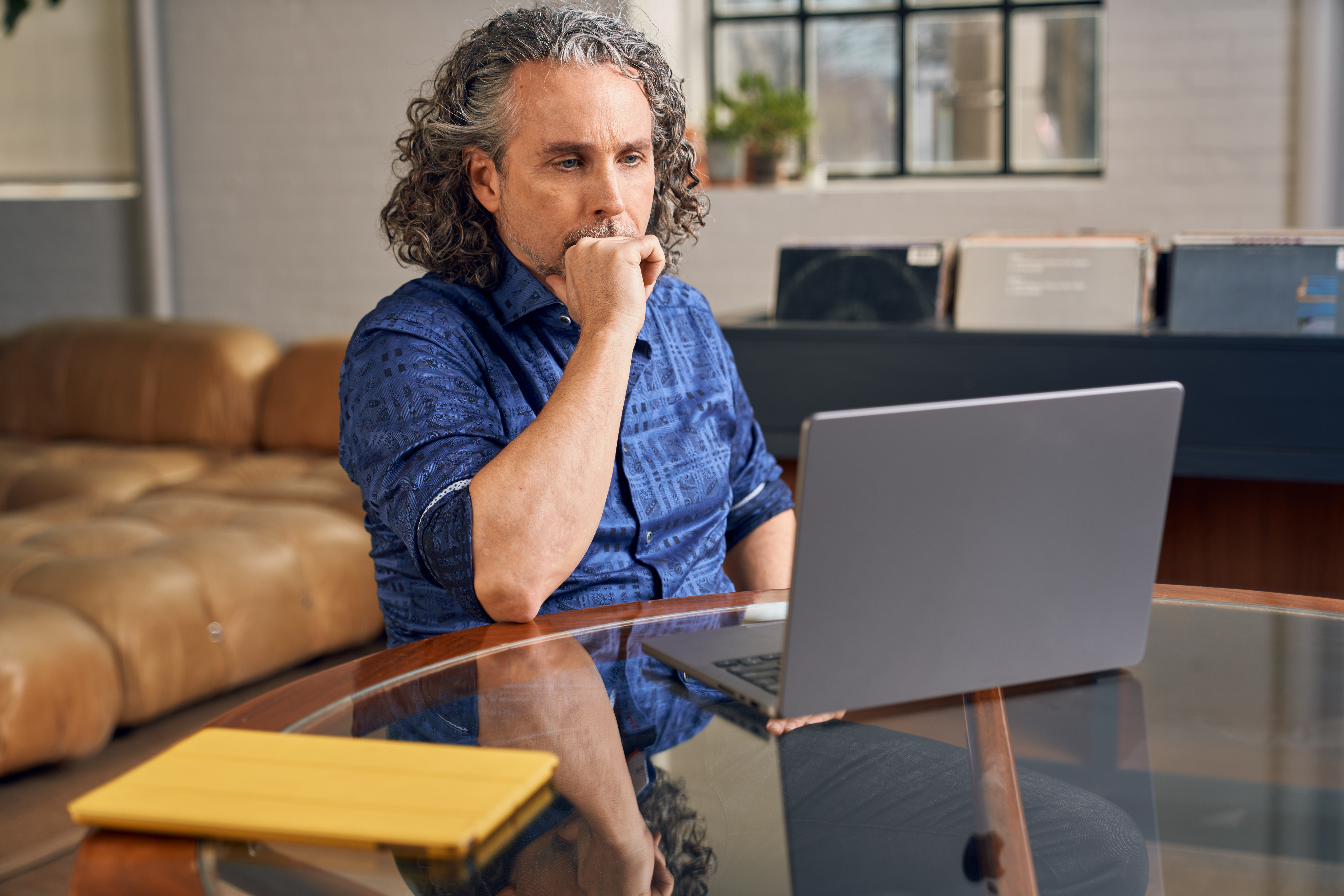 A man sat concentrating at his laptop on a glass table at home, wearing a blue shirt.