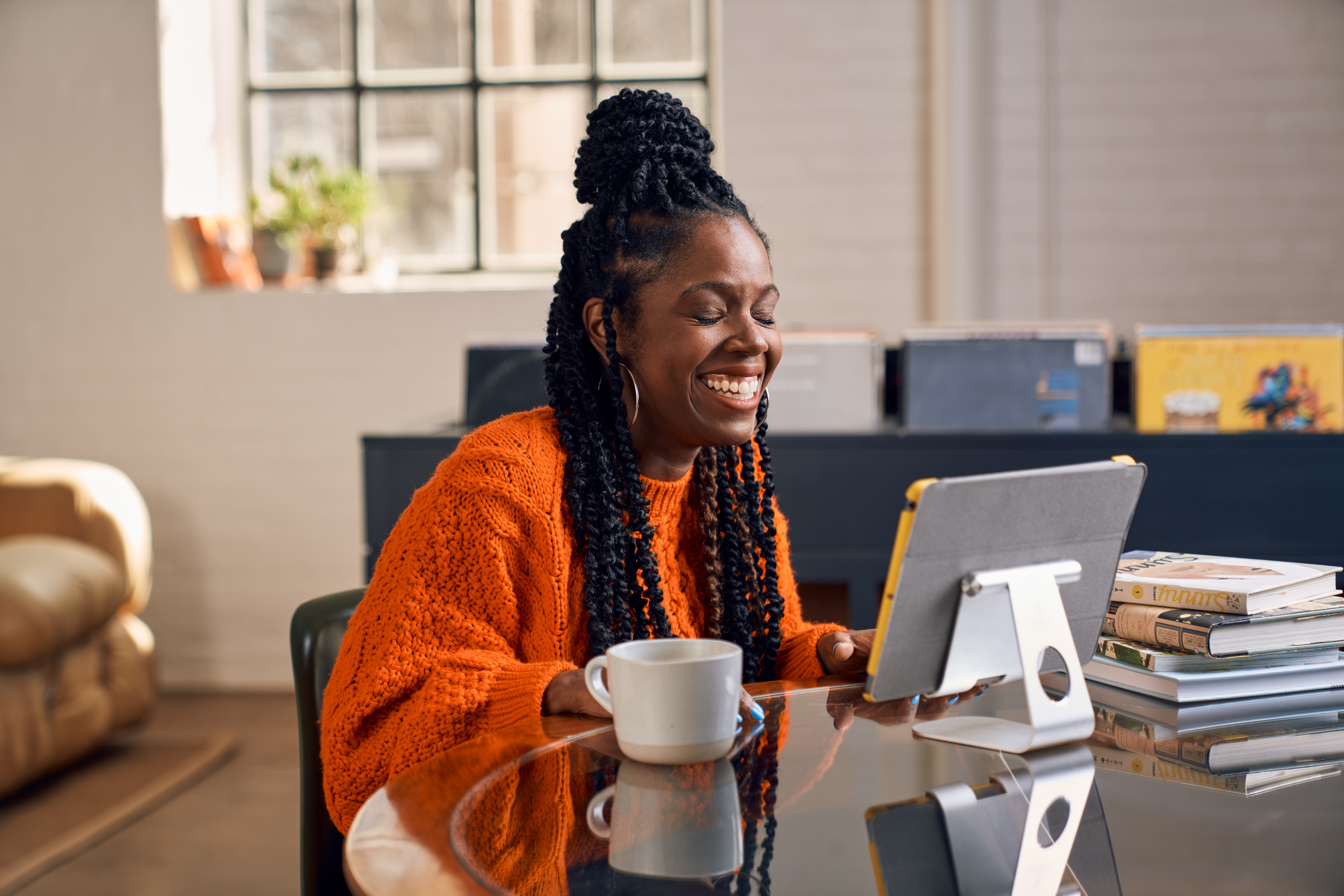 Woman sat at her table in an orange jumper, smiling and working at a tablet.