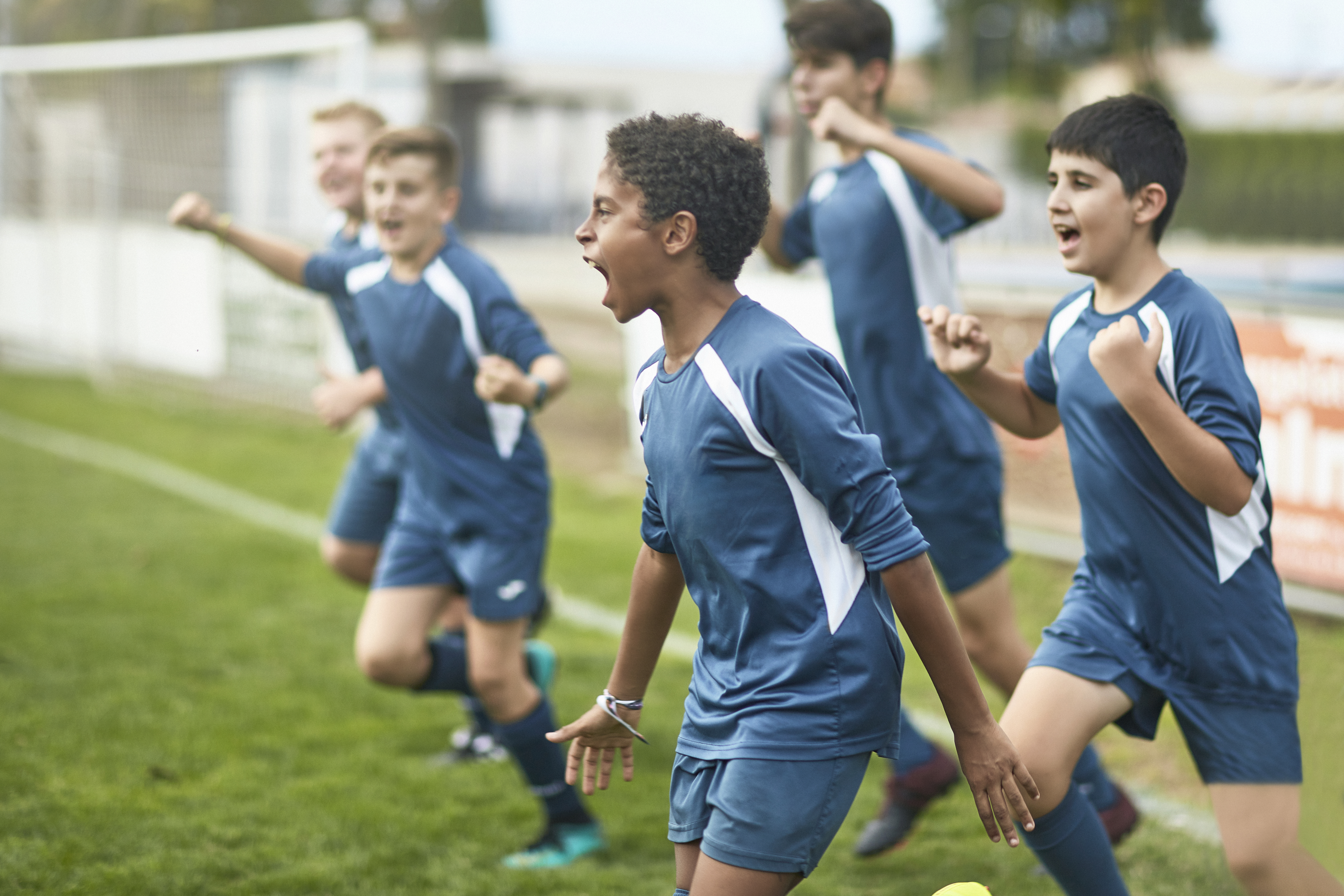 Energetic preteen and teenage male footballers cheering and punching the air as they run onto field for training session. Sports. Outside.