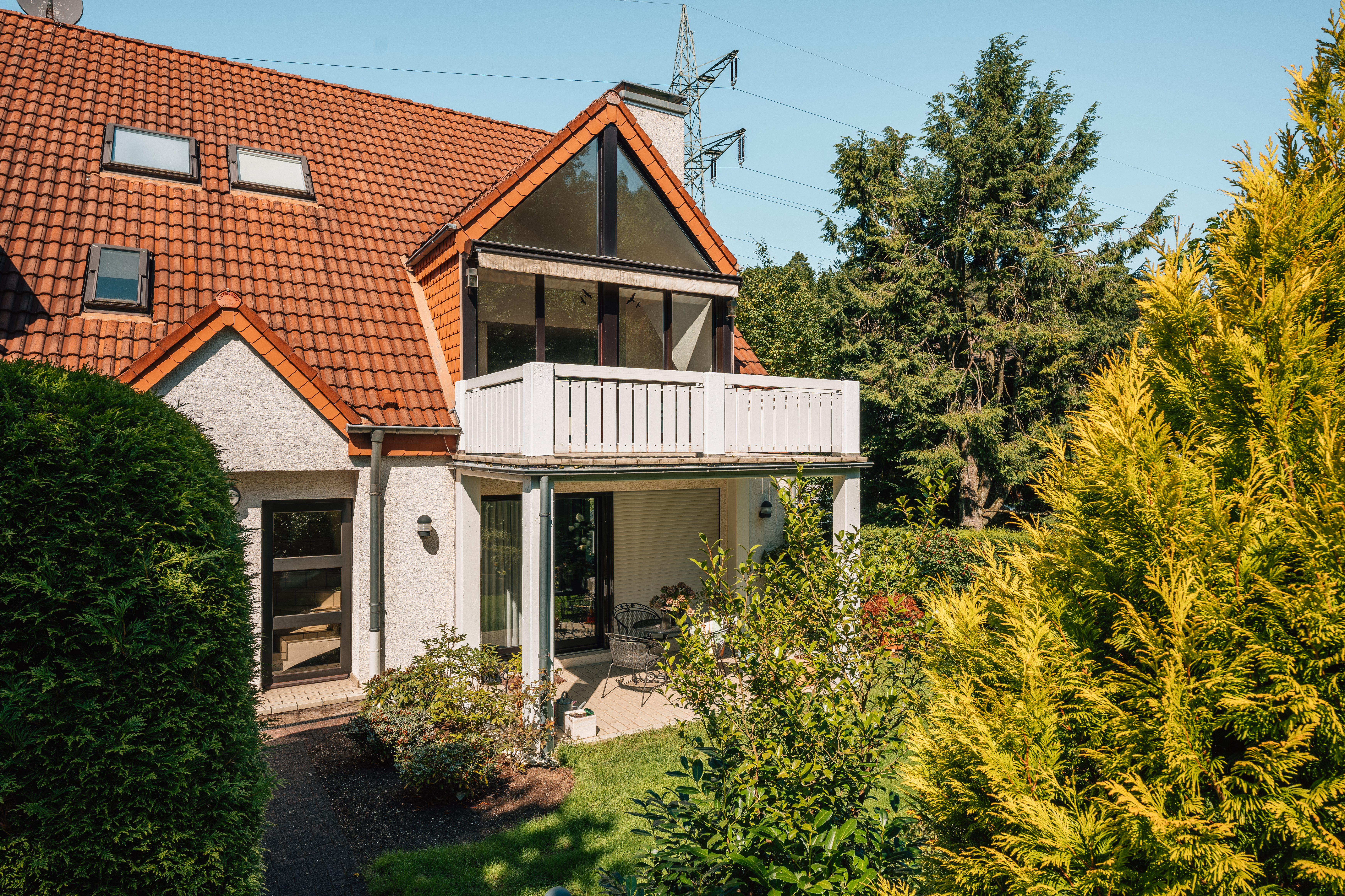 Außenansicht eines Hauses mit Balkon, Terrasse und Garten, umgeben von Bäumen und Sträuchern.