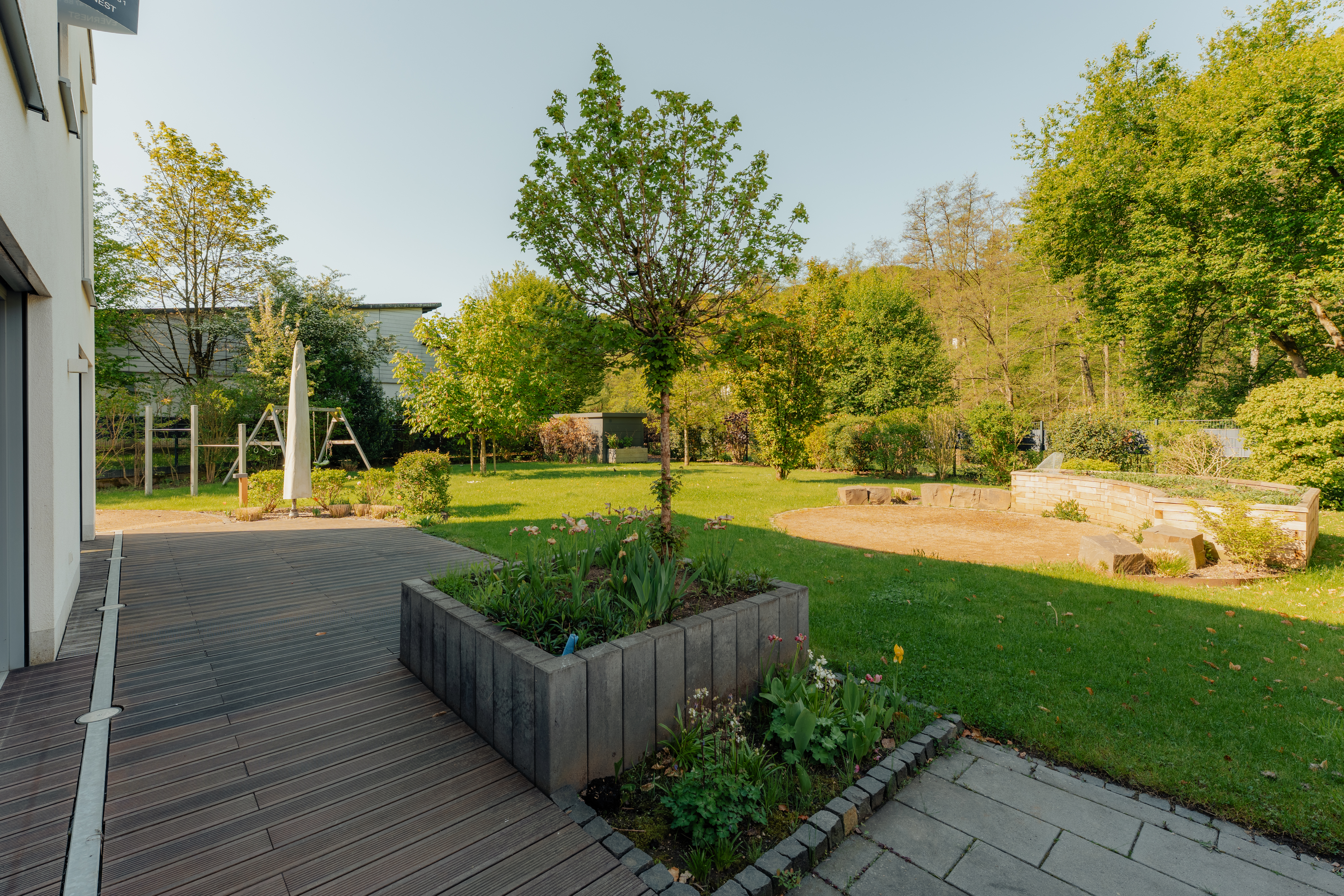 Grüner Garten mit Rasenfläche, Bäumen, Sandfläche und Hochbeet neben einer Terrasse.