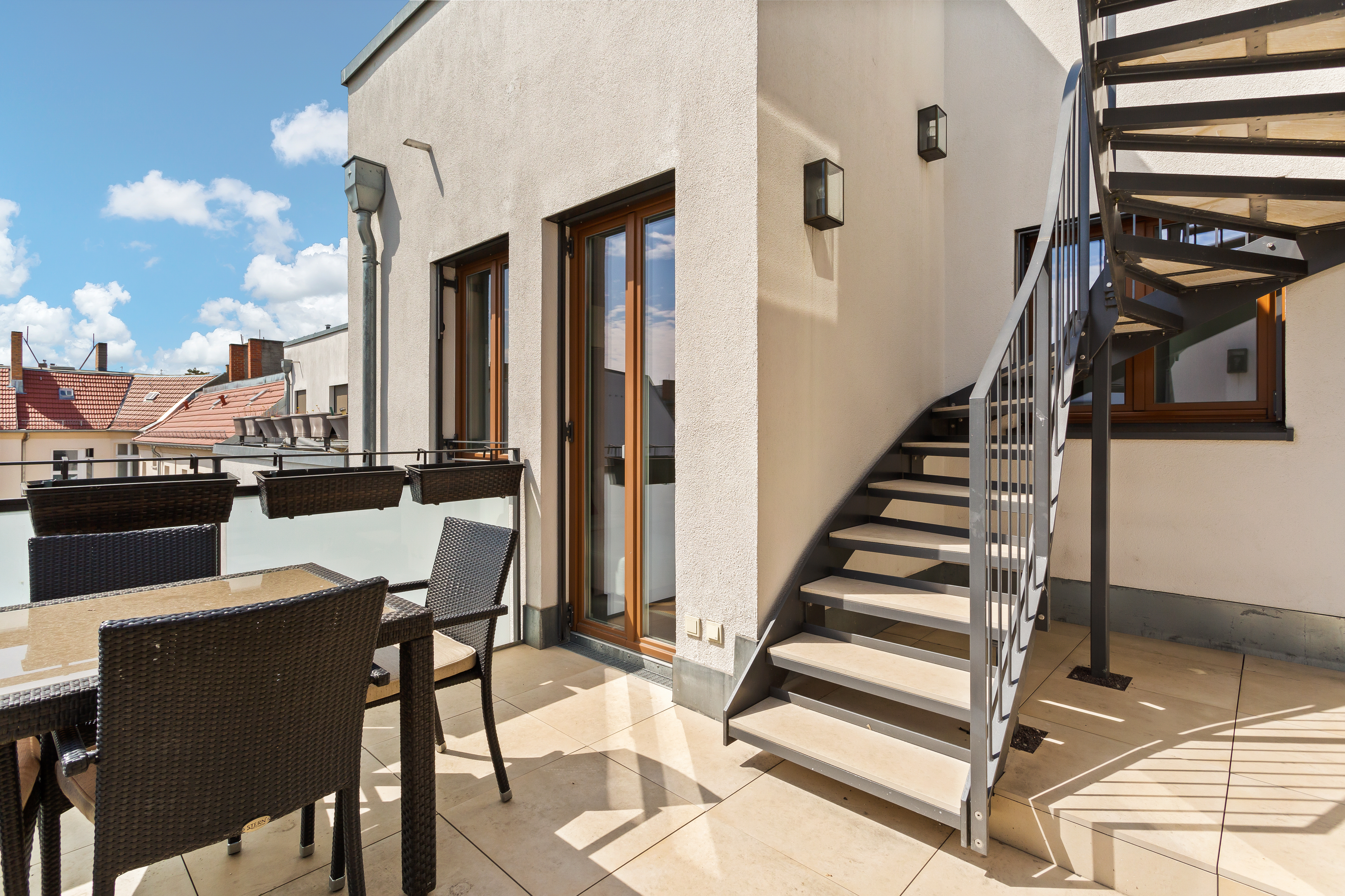 Möblierter Balkon mit Glastisch, Stühlen und einer Wendeltreppe an einer modernen Fassade.