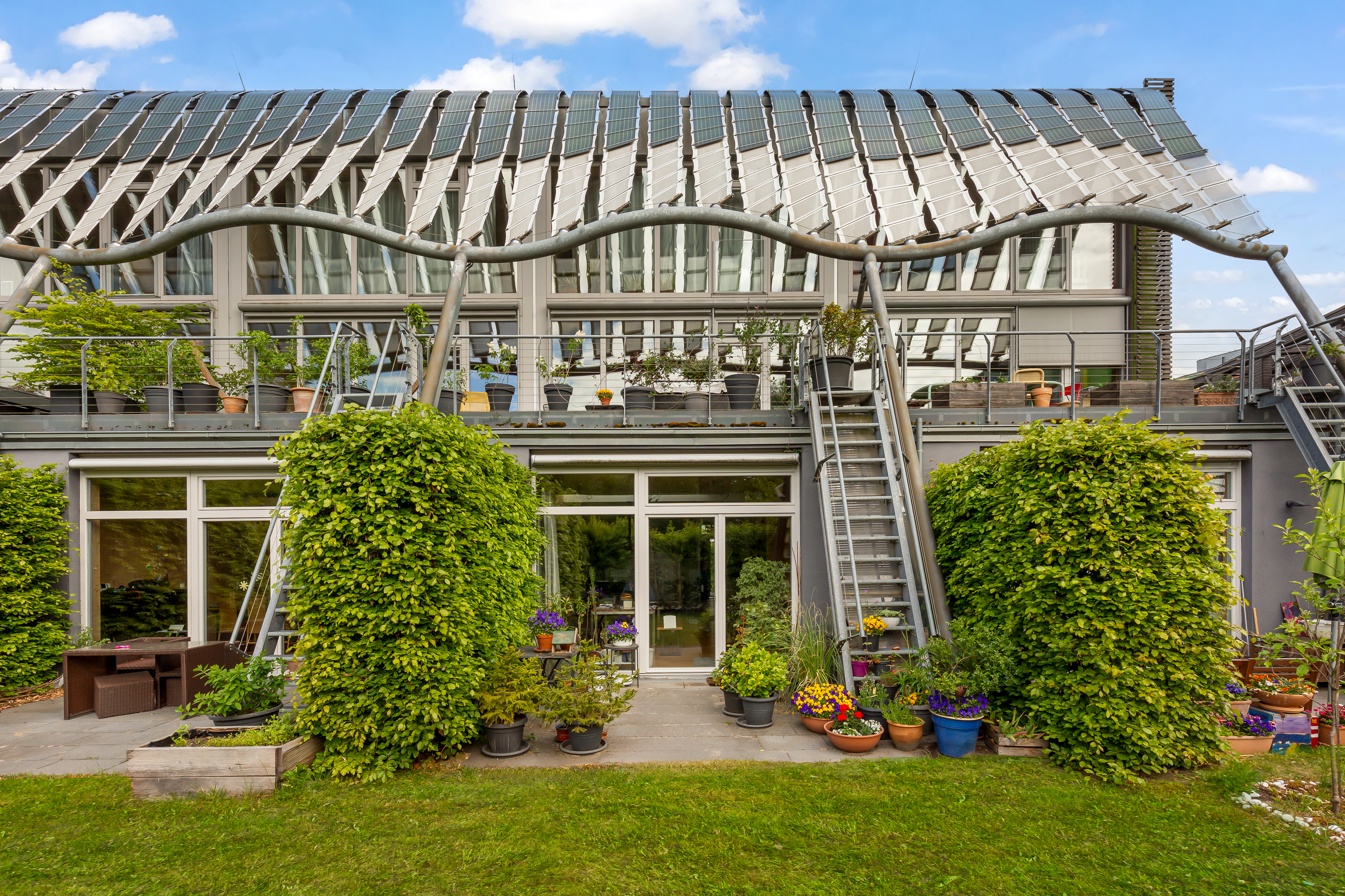 Moderne Hausfassade mit großen Fenstern, Balkon, Außentreppe und vielen Pflanzen in Töpfen.