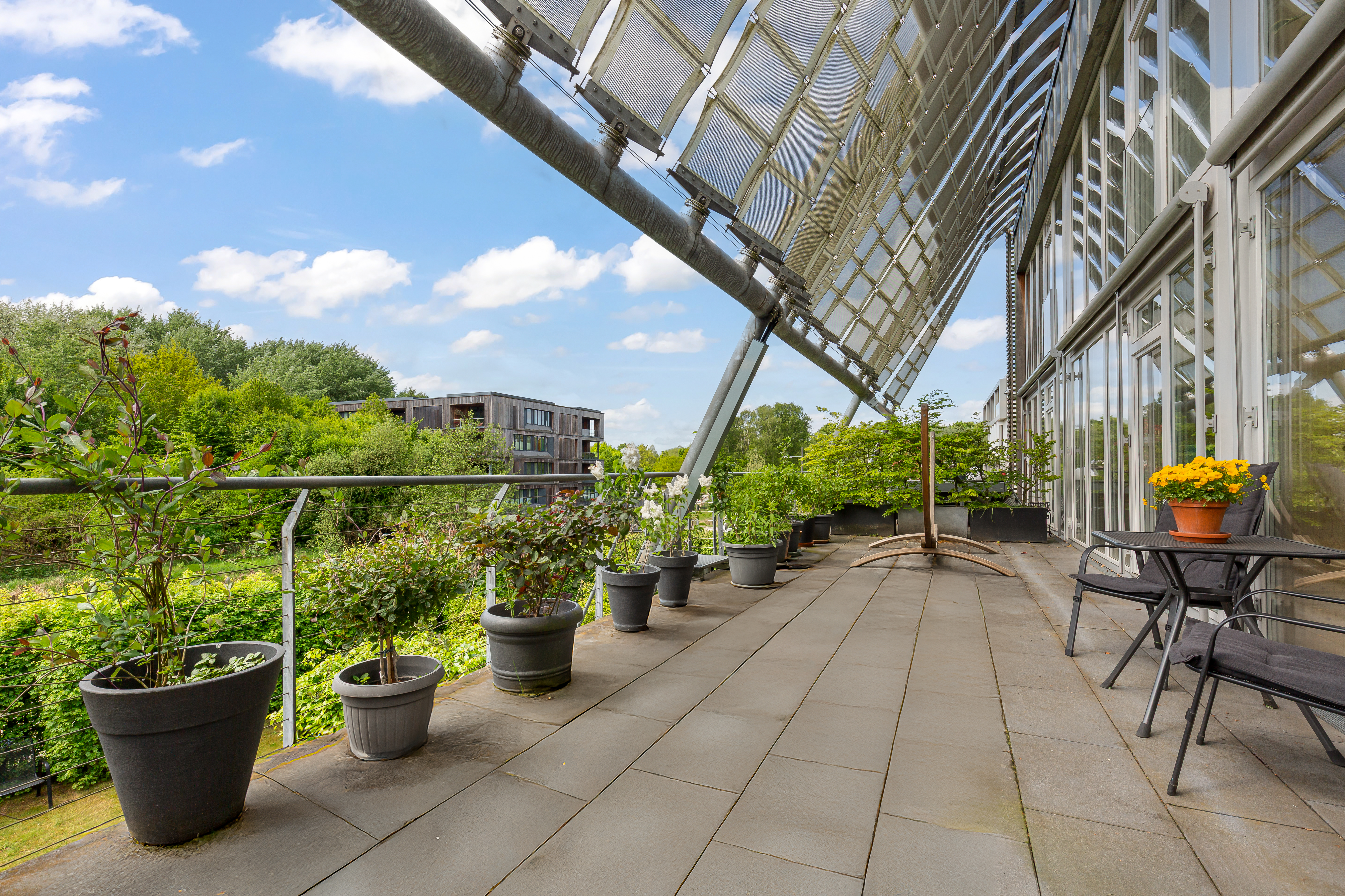 Große Terrasse mit Blumentöpfen, Stühlen, Tisch und Glasfassade, Blick ins Grüne.