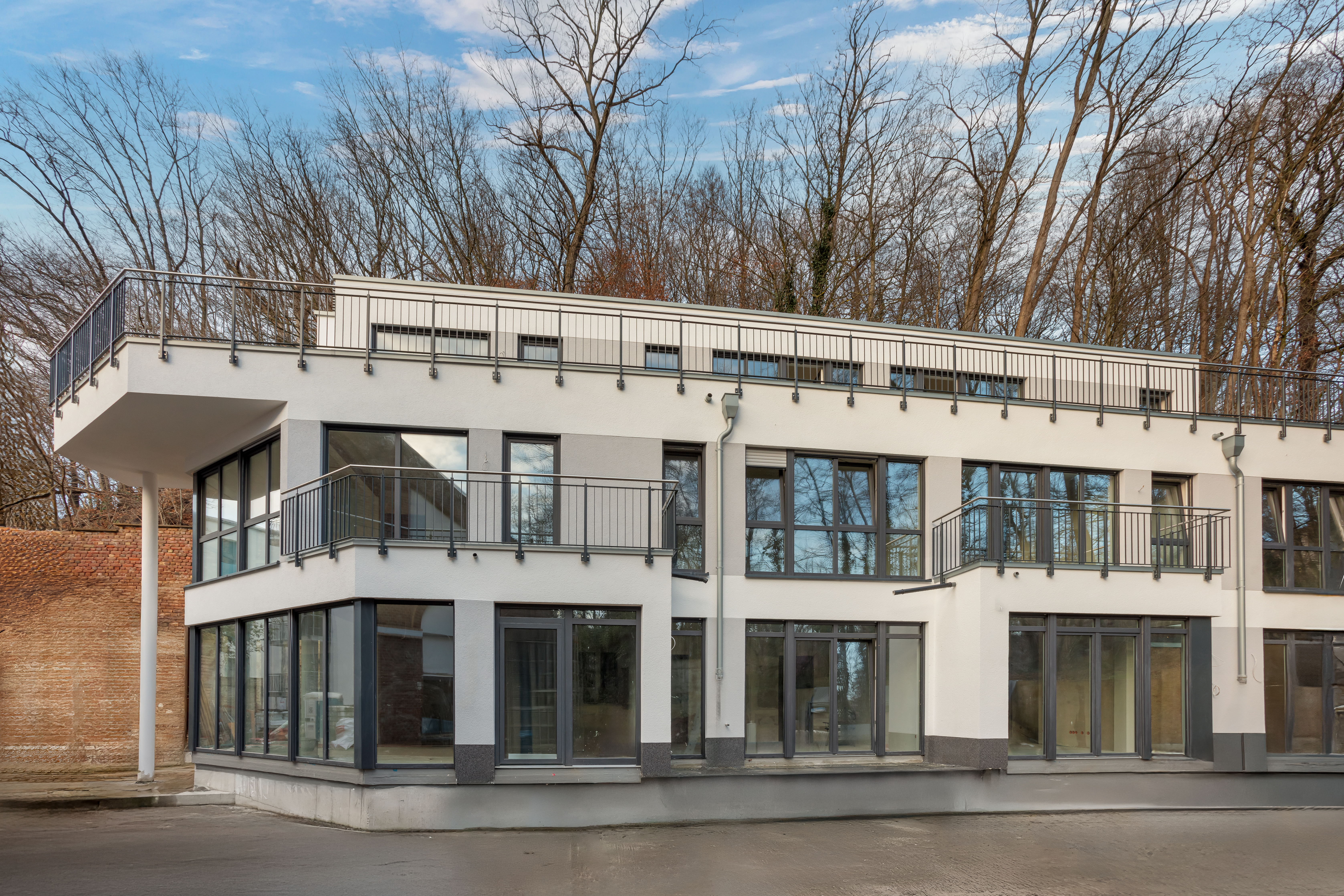 Modernes Wohngebäude mit großen Fenstern, Balkonen und flachem Dach vor Bäumen.