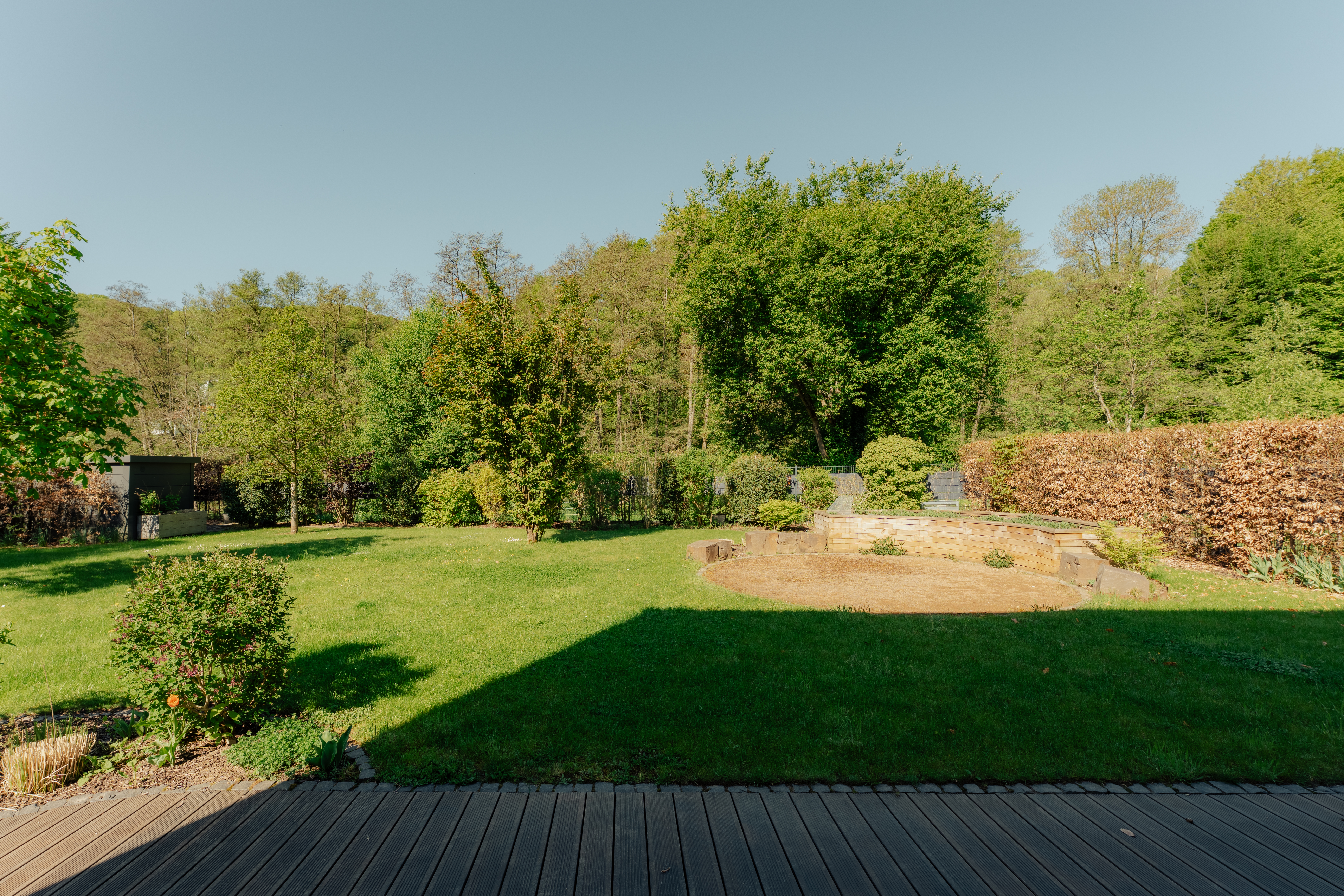 Großer Garten mit Rasenfläche, Büschen, Bäumen und Holzboden im Vordergrund.