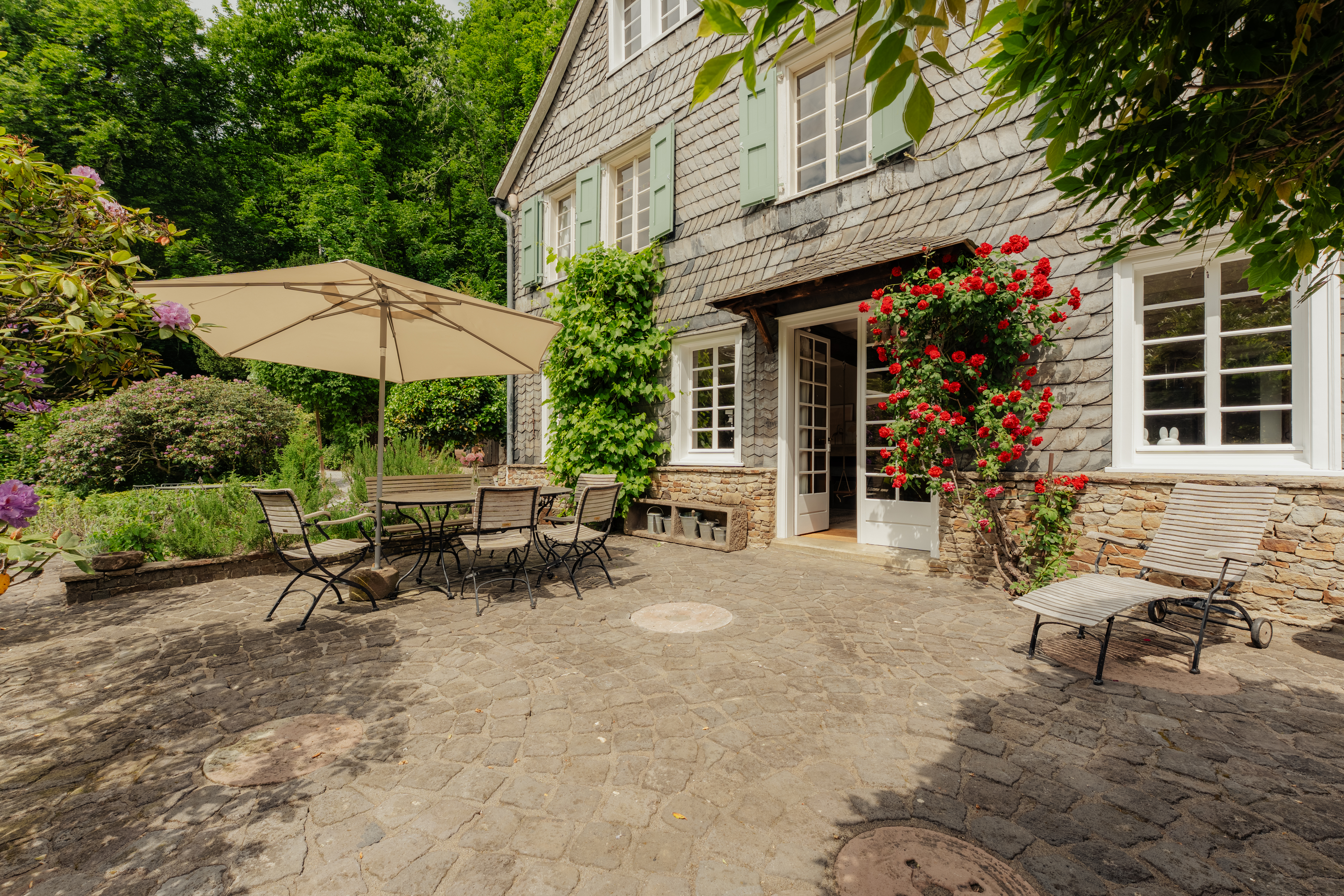 Gepflasterte Terrasse mit Tisch, Stühlen, Sonnenschirm und blühenden Rosen an einem Haus.