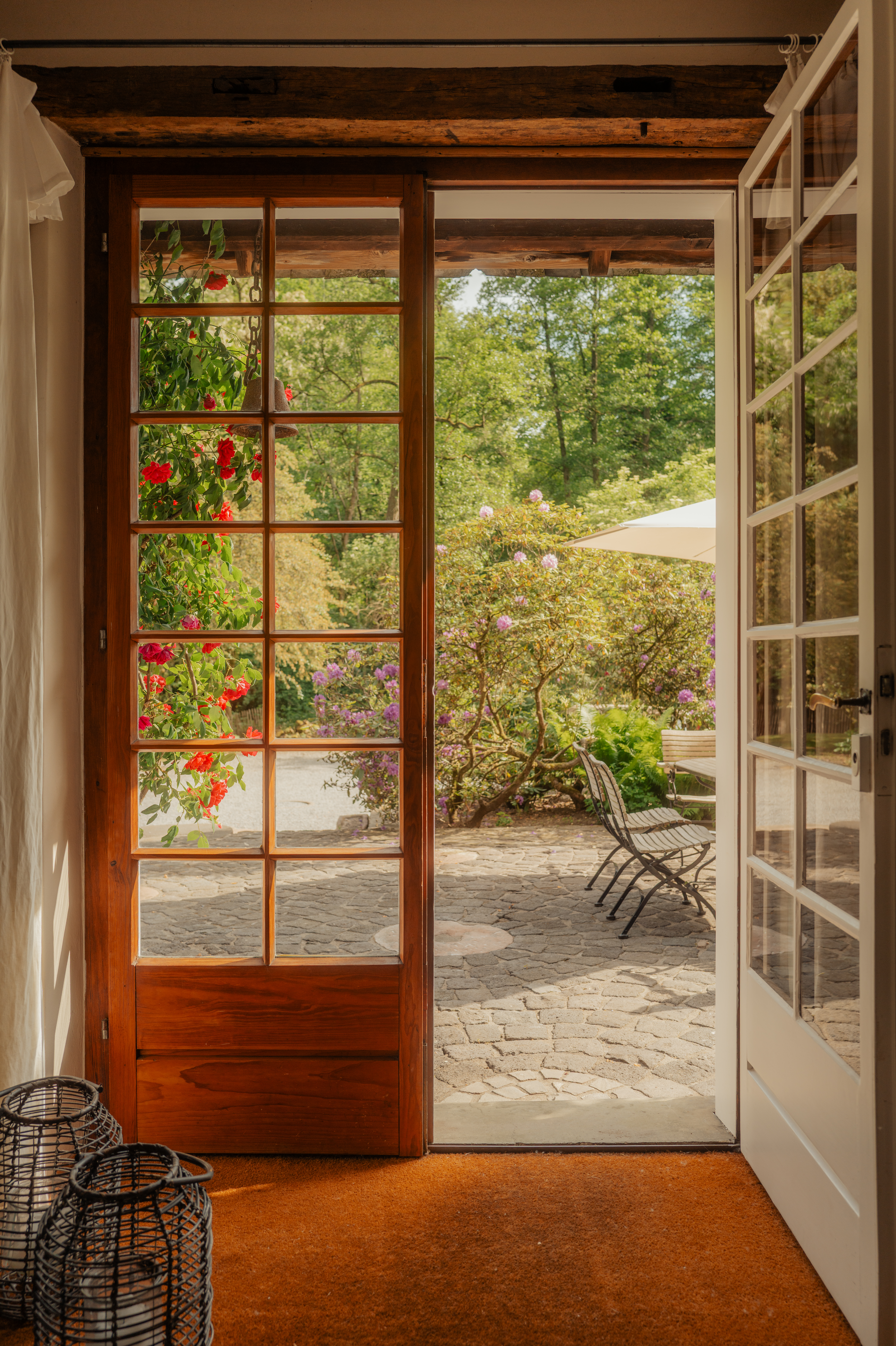 Offene Glastür mit Blick auf eine Terrasse mit Garten, Blumen und Sitzmöbeln.