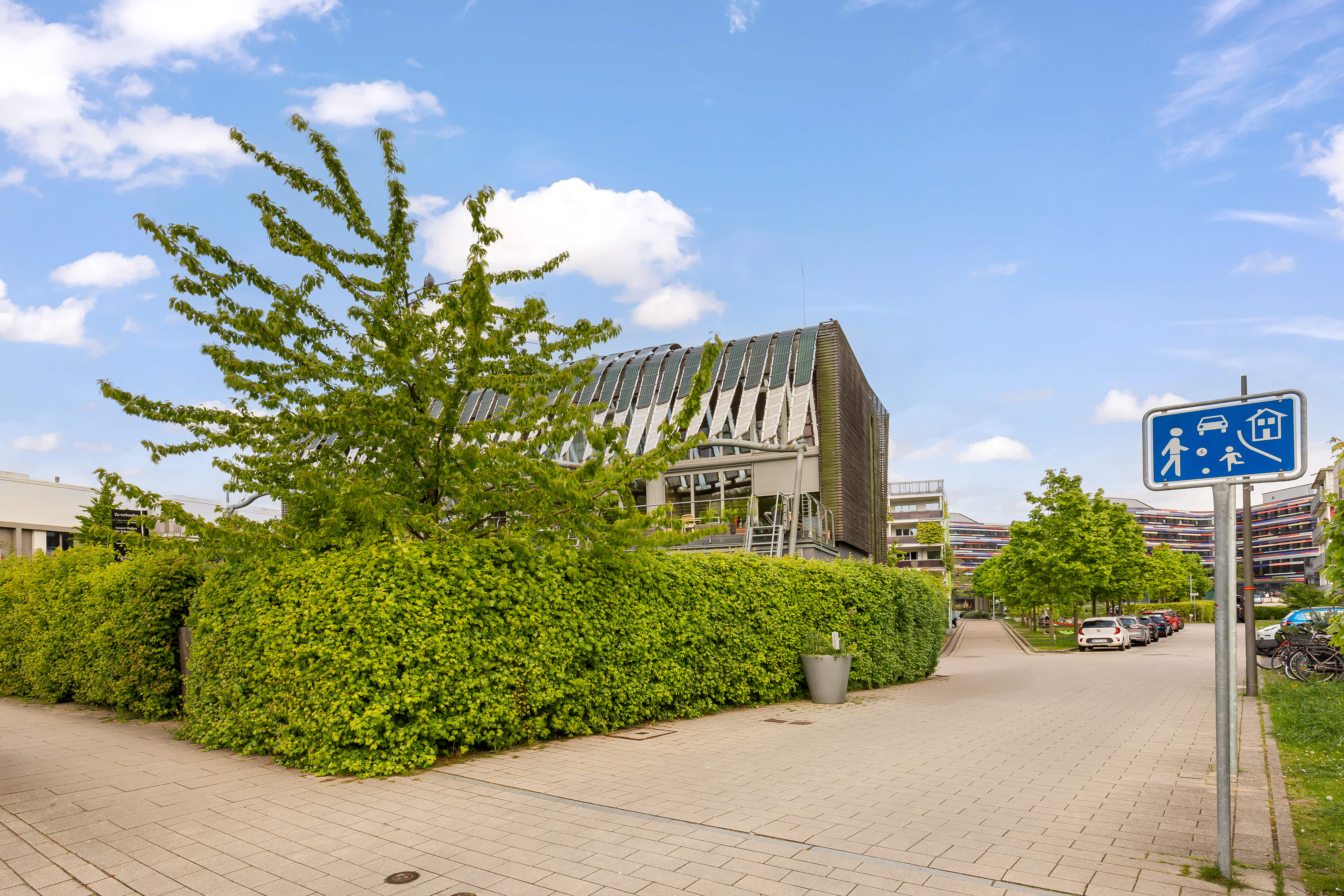 Moderne Wohngebäude mit Glasdach, viel Grün und Gehweg, daneben Verkehrsschild.