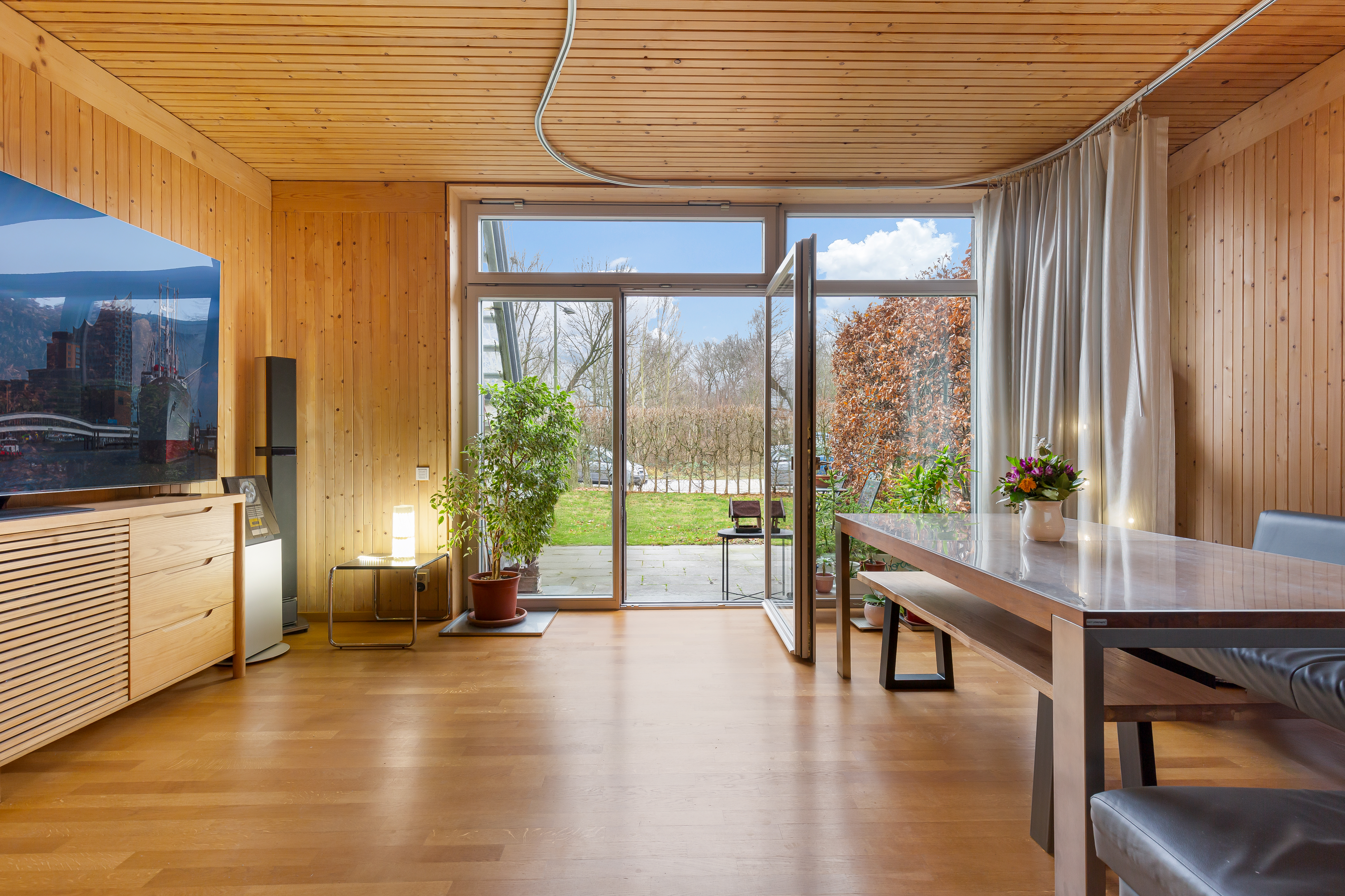 Helles Wohnzimmer mit Holzwänden, großem Fenster, Esstisch und Pflanzen, Blick in den Garten.