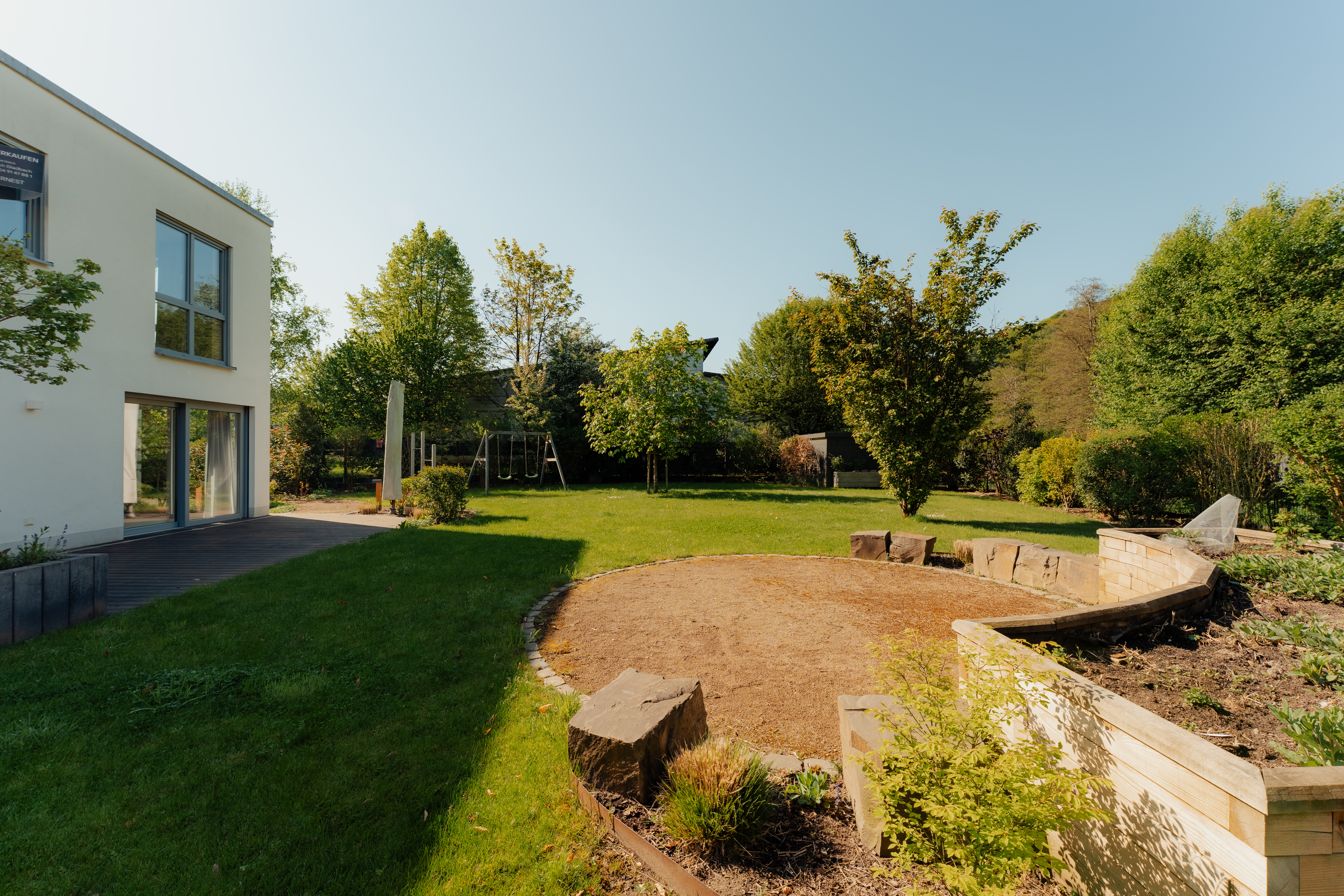 Moderner Garten mit Rasen, Bäumen, Sandfläche, Schaukel und Terrasse an einem Wohnhaus.