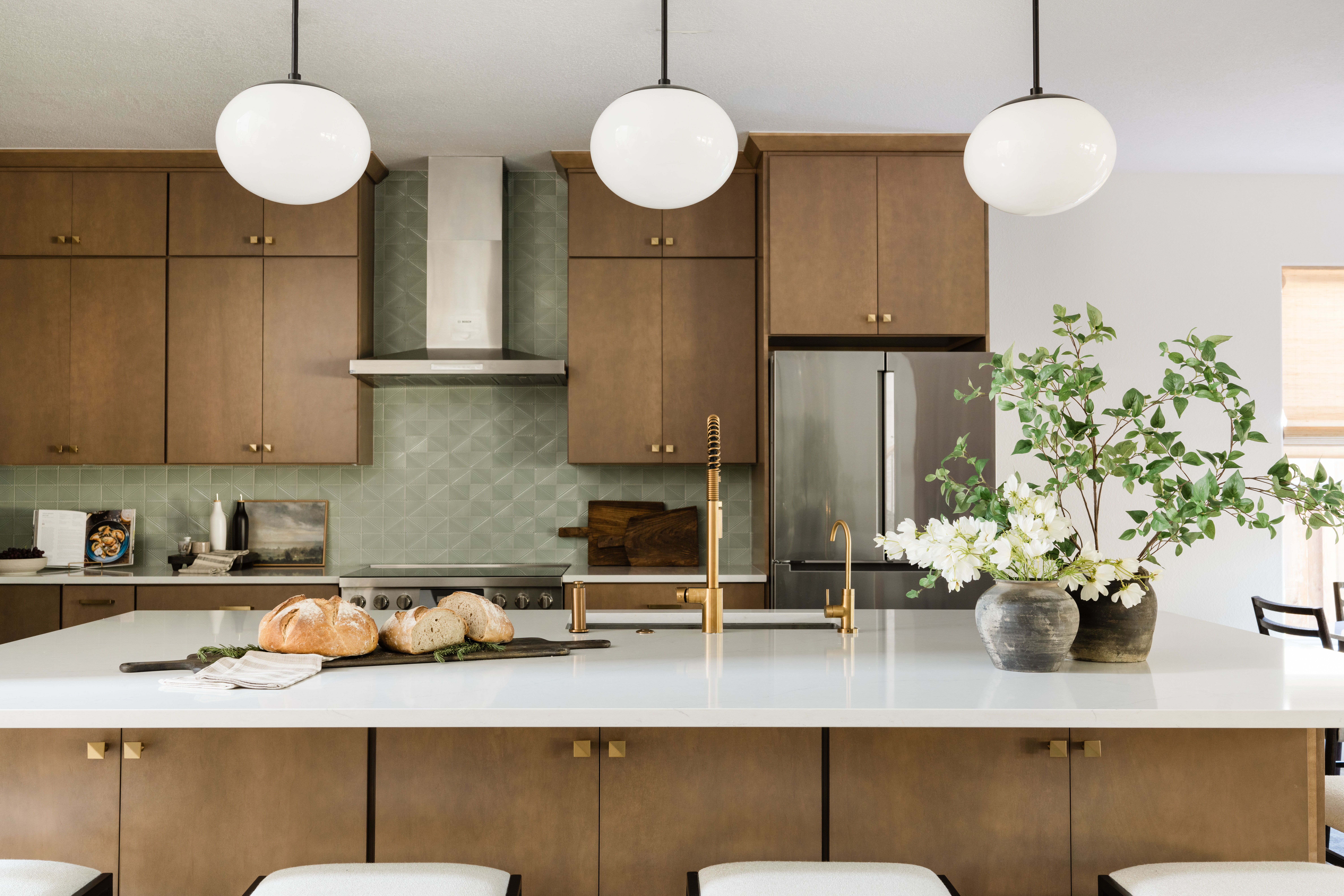 An image of a kitchen, zoomed in on the kitchen island, centered in the room. The kitchen has white counter tops, natural wood colored cabinets, and a sage green tiled backsplash. Three globe pendant light hang over the kitchen island. Vases with flowers and greenery are displayed on top of the island on the right side with a wooden board with loaves of artisan breads on the left. 