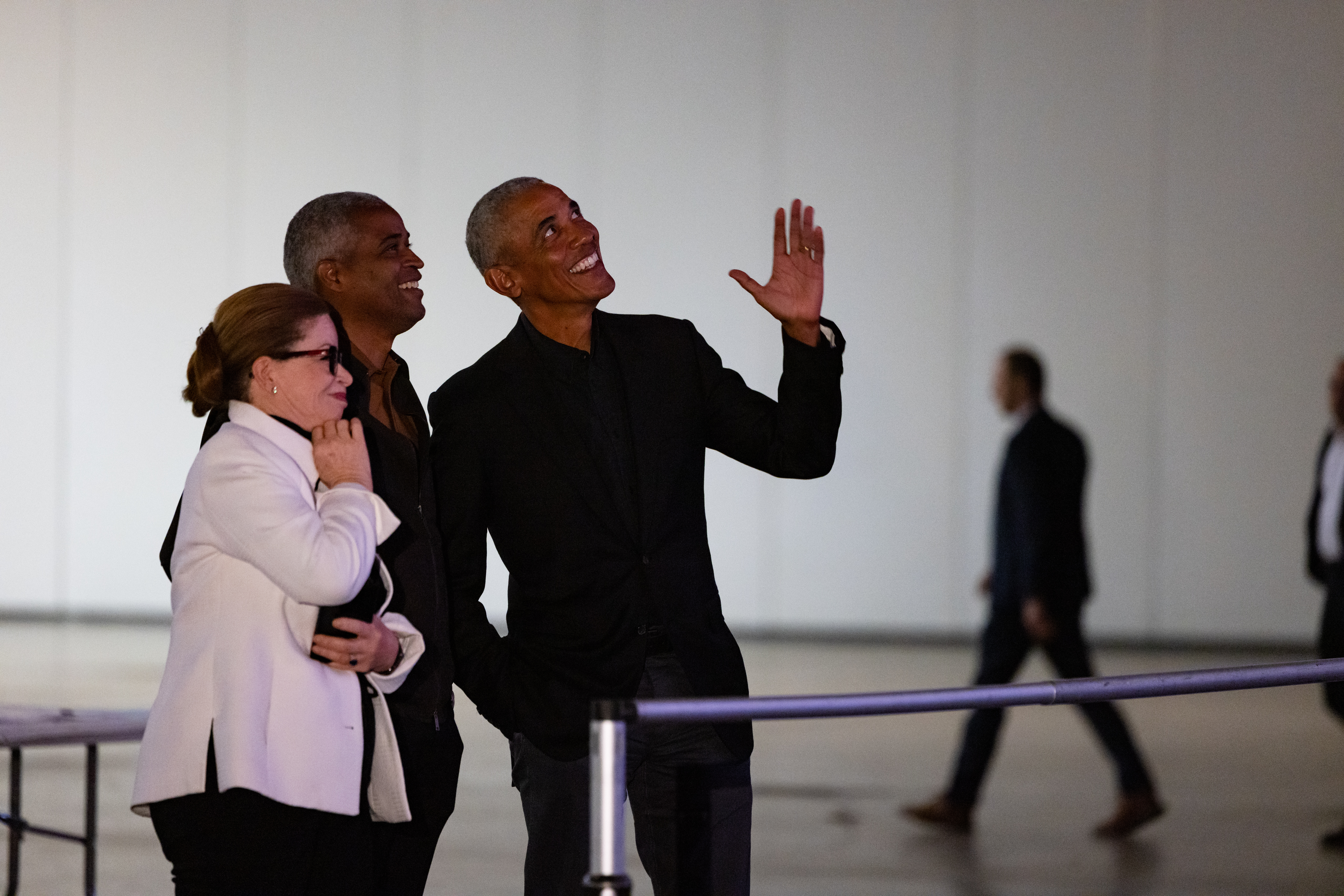 The image is a photograph of Barack Obama, Valerie Jarrett, and Marty Nesbitt looking at the Power of Words prototype. Barack Obama stands to the far left and is gesturing towards the Power of Words prototype with his left hand. He is wearing a black blazer, a black shirt, and dark denim pants. Marty Nesbitt stands in the middle: he is smiling. He has a brown dark complexion and gray hair. Valerie Jarrett stands on the far right. She is smiling with no teeth. Her hair is pulled back into a chingon and she is wearing black glasses. She has her right hand to her mouth. She is wearing a white blazer and black pants. Behind Valerie Jarrett, Marty Nesbitt, and Barack Obama is a white wall.  