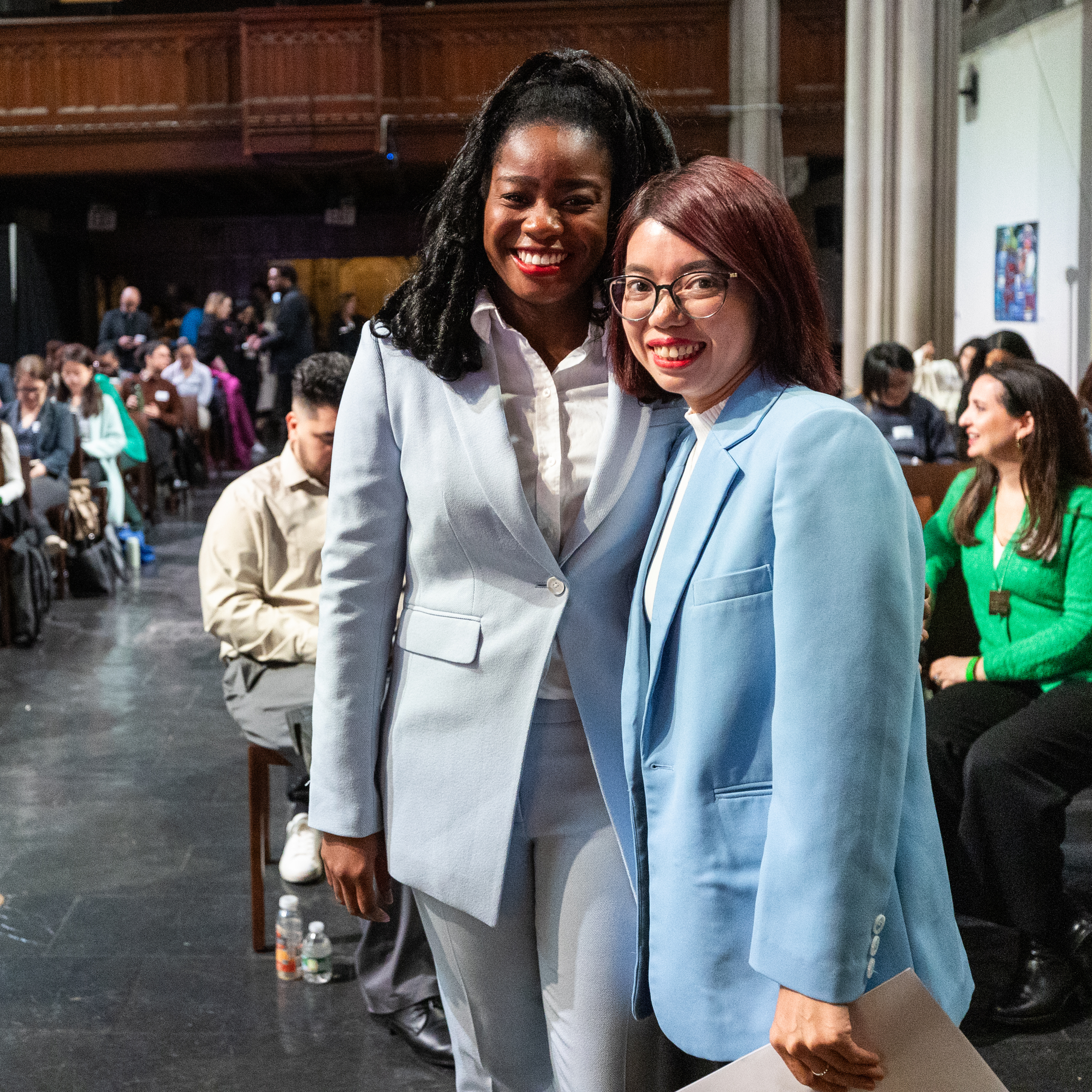 Amanda Morrall, a Black woman with a dark skin tone, and Huong Dang, a woman with a light skin tone pose in front of an audience of people with a range of ages and light to deep skin tones. Both women are wearing powder blue pantsuits, white blouses, and red lipstick. 
