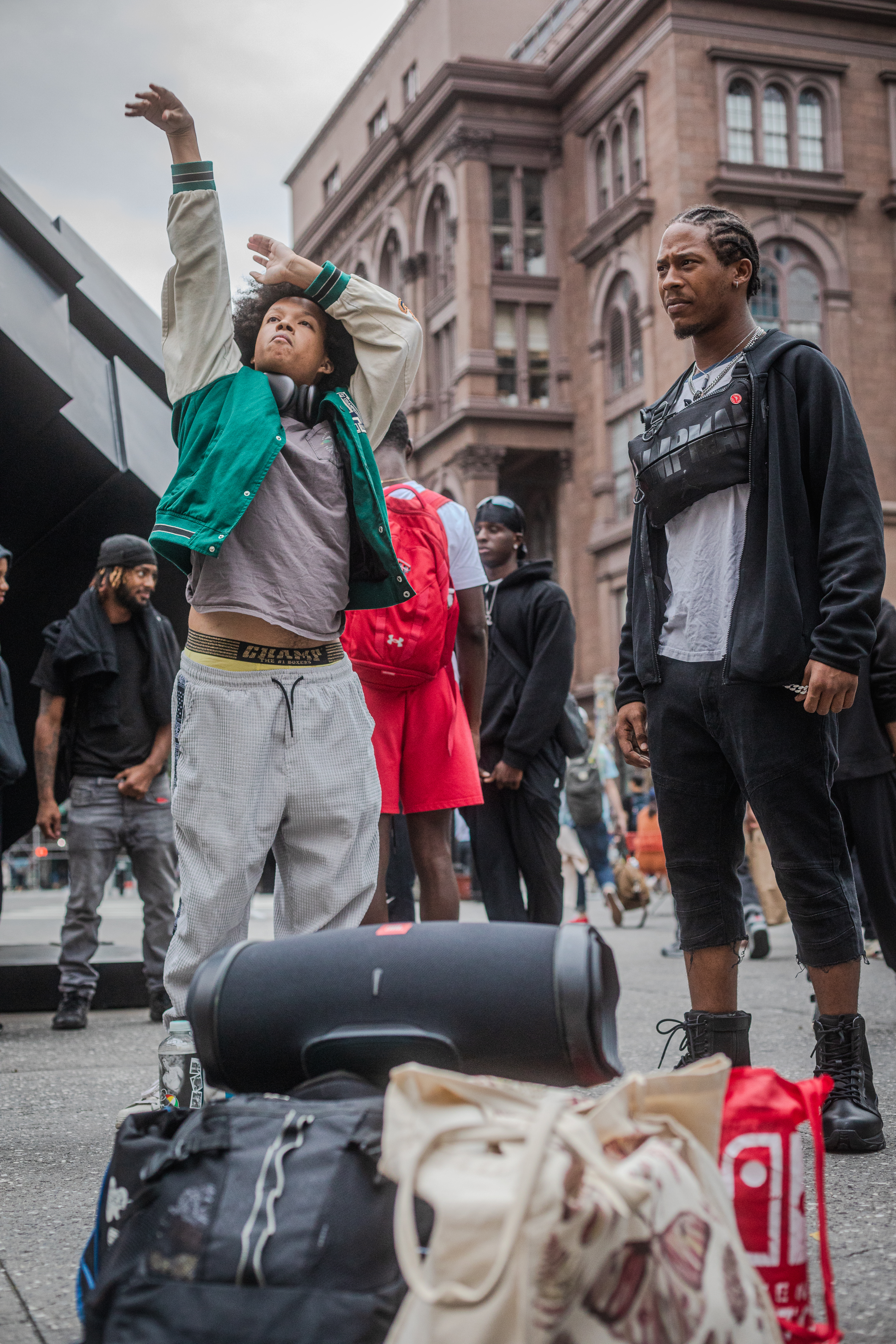 The image is a photo of several men on a street outside. Behind the men is a brown stone building. To the left of the men is an abstract statue. There are several men standing in a circle in the foreground of the image. In the front of the image are two men. The first man is standing in a position to symbolize throwing a basketball shot. He has a light deep medium complexion, a large black afro and is wearing a green and beige lettermans jacket, a grey shirt, and grey sweatpants. The second man has a medium deep complexion, a goatee, and cornrows in his hair. He is wearing a black hoodie, a grey shirt, cut off black pants cut to the calf, and black combat boots. He has a fanny pack sitting on his neck. In front of the men are several backpacks sitting on the ground.