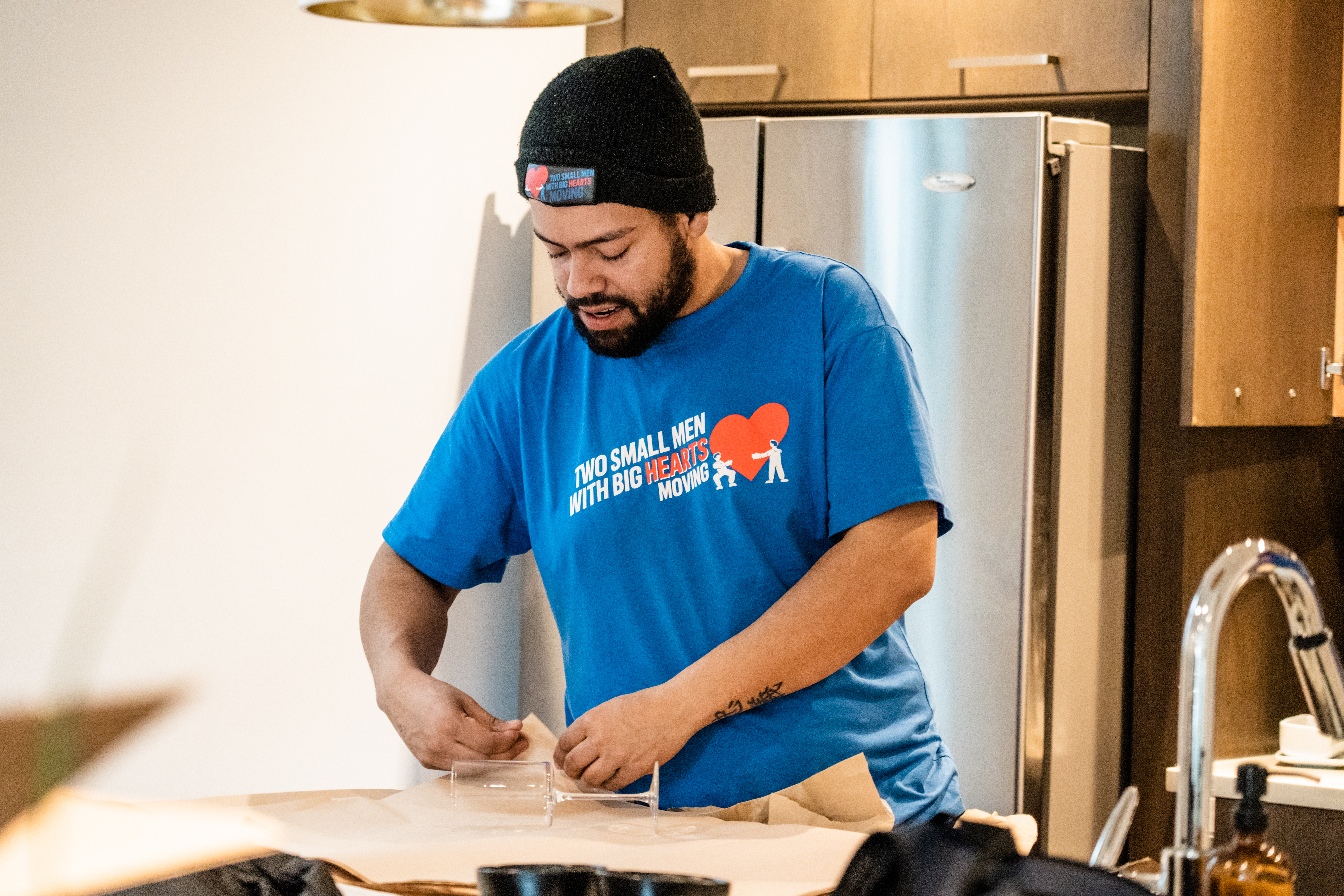 Man in a Two Small Men with Big Hearts Moving shirt packing glassware in a kitchen.