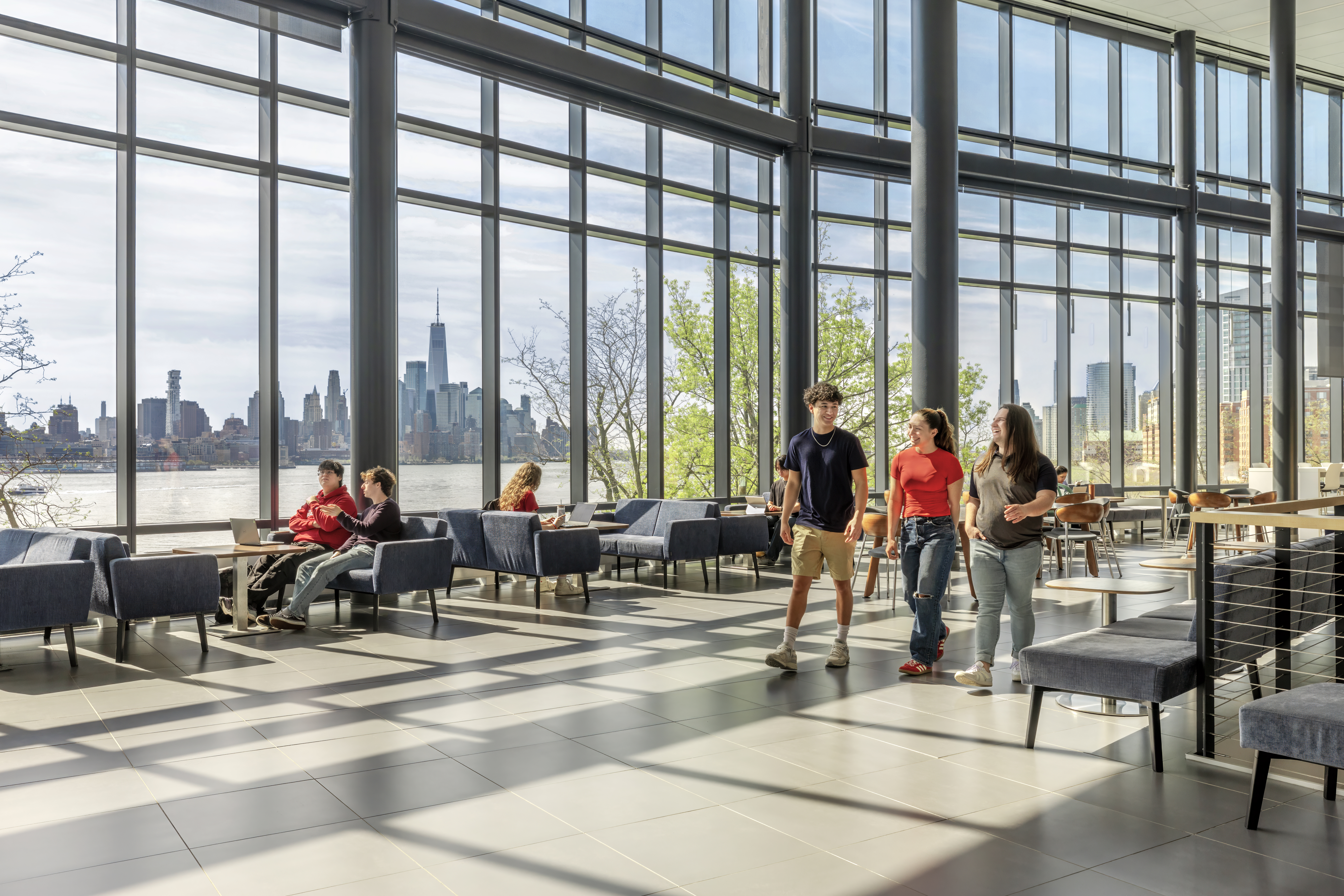 Students walk inside the University Center Complex on a sunny day, the NYC skyline in the background.