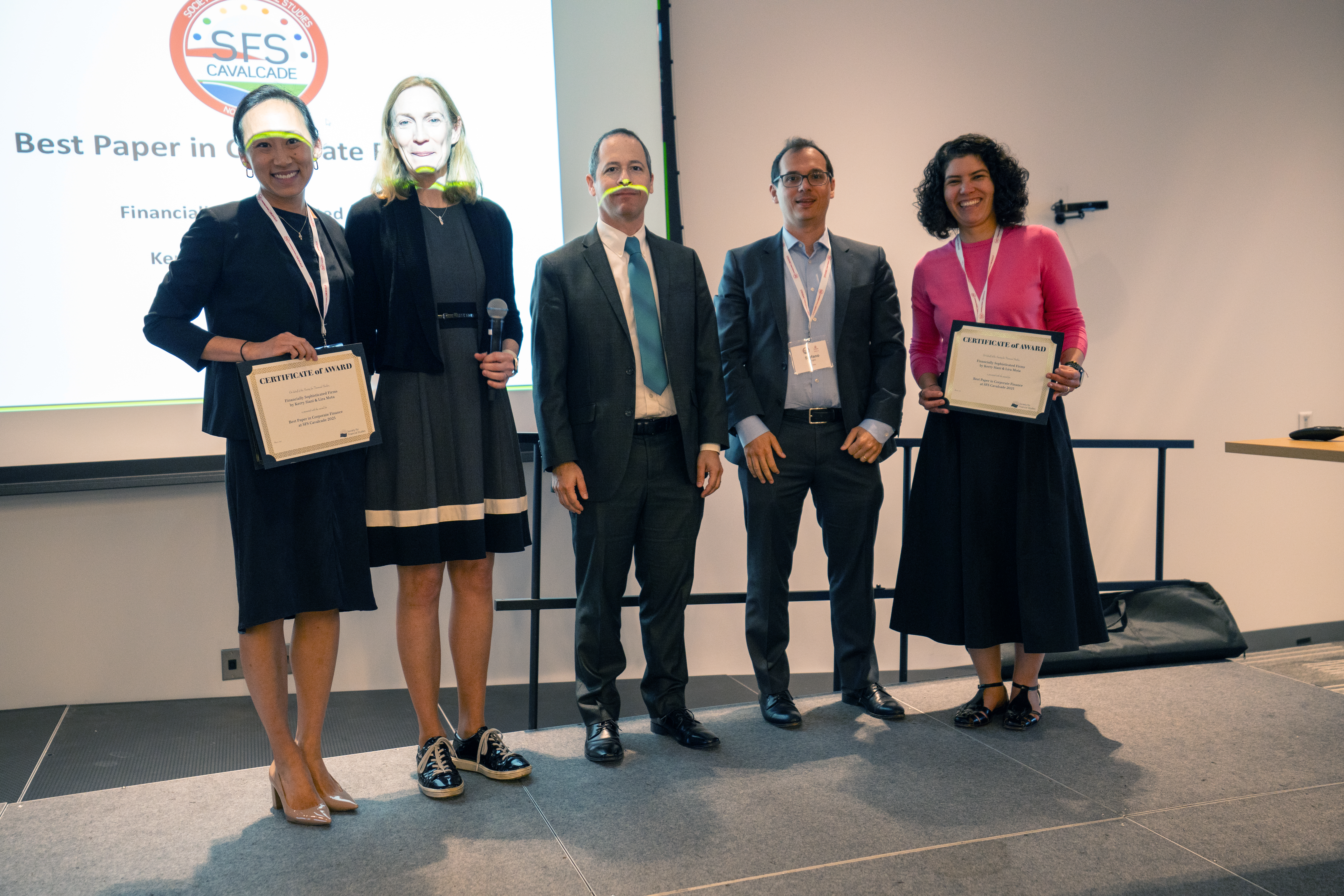 Five professionals pose at an awards ceremony, with two women holding certificates flanking three men in suits. Behind them is a projection screen showing "Best Paper in Corporate Finance" with an SFS logo in a modern conference room.