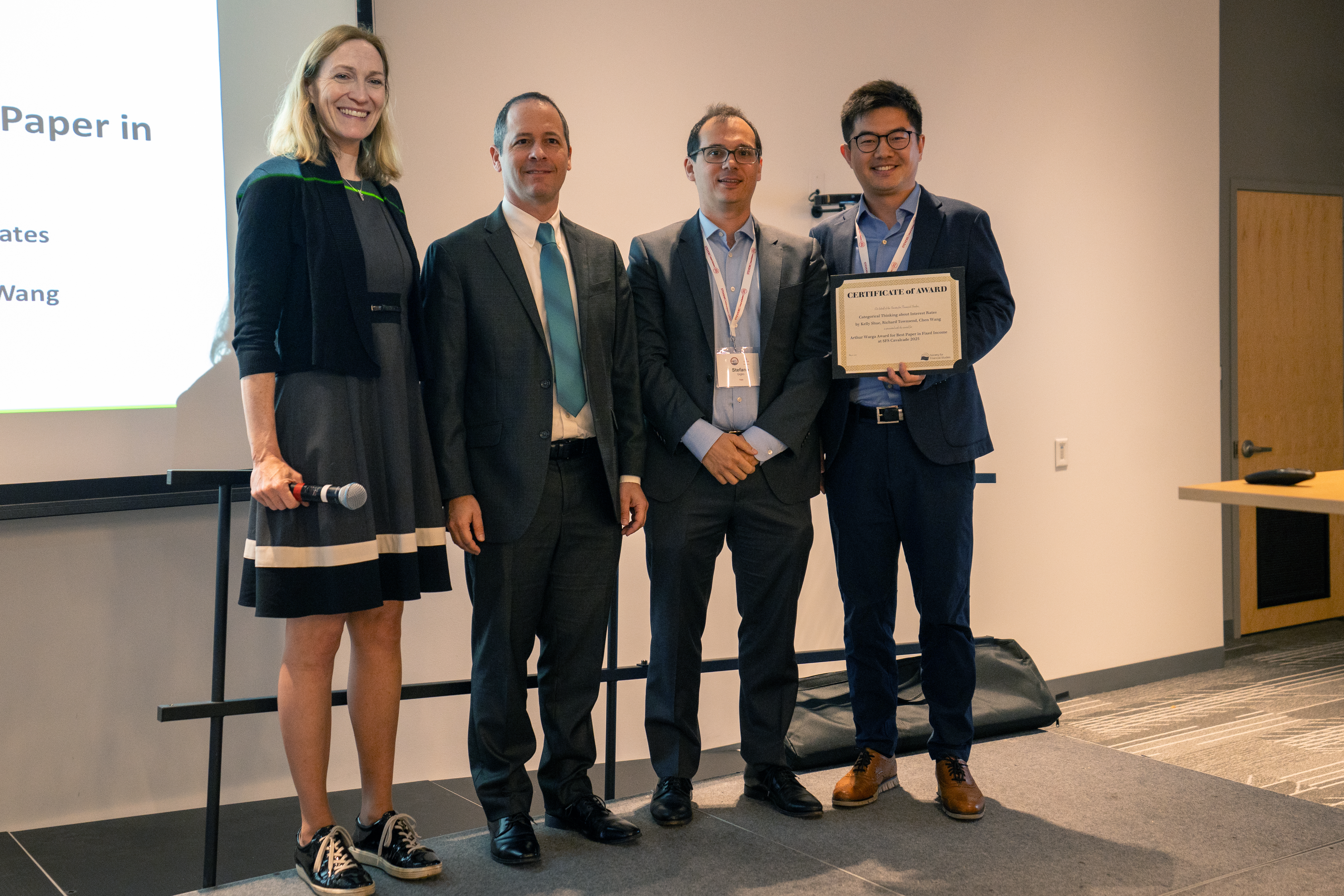 Four professionals pose at an academic award ceremony. A woman in a striped dress holds a microphone while three men in business suits stand beside her, with one holding a certificate, against a projection screen in a conference room.