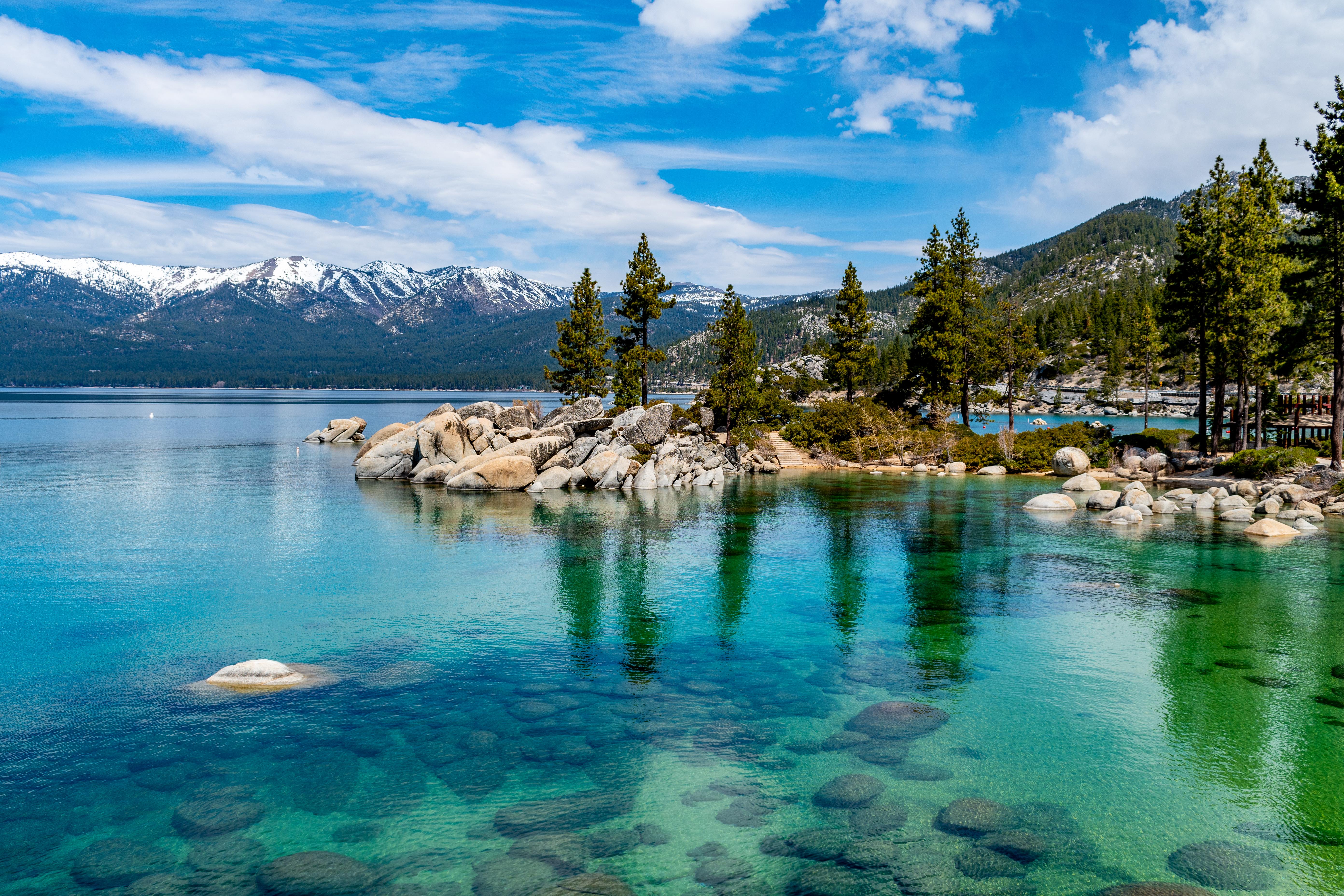 A serene view of Lake Tahoe, CA, with crystal-clear turquoise waters, smooth rocks, and pine trees along the shoreline. Snow-capped mountains rise in the background, under a bright blue sky with scattered clouds. This image perfectly encapsulates the tranquil and breathtaking natural beauty often found near luxury homes with private access to natural wonders.