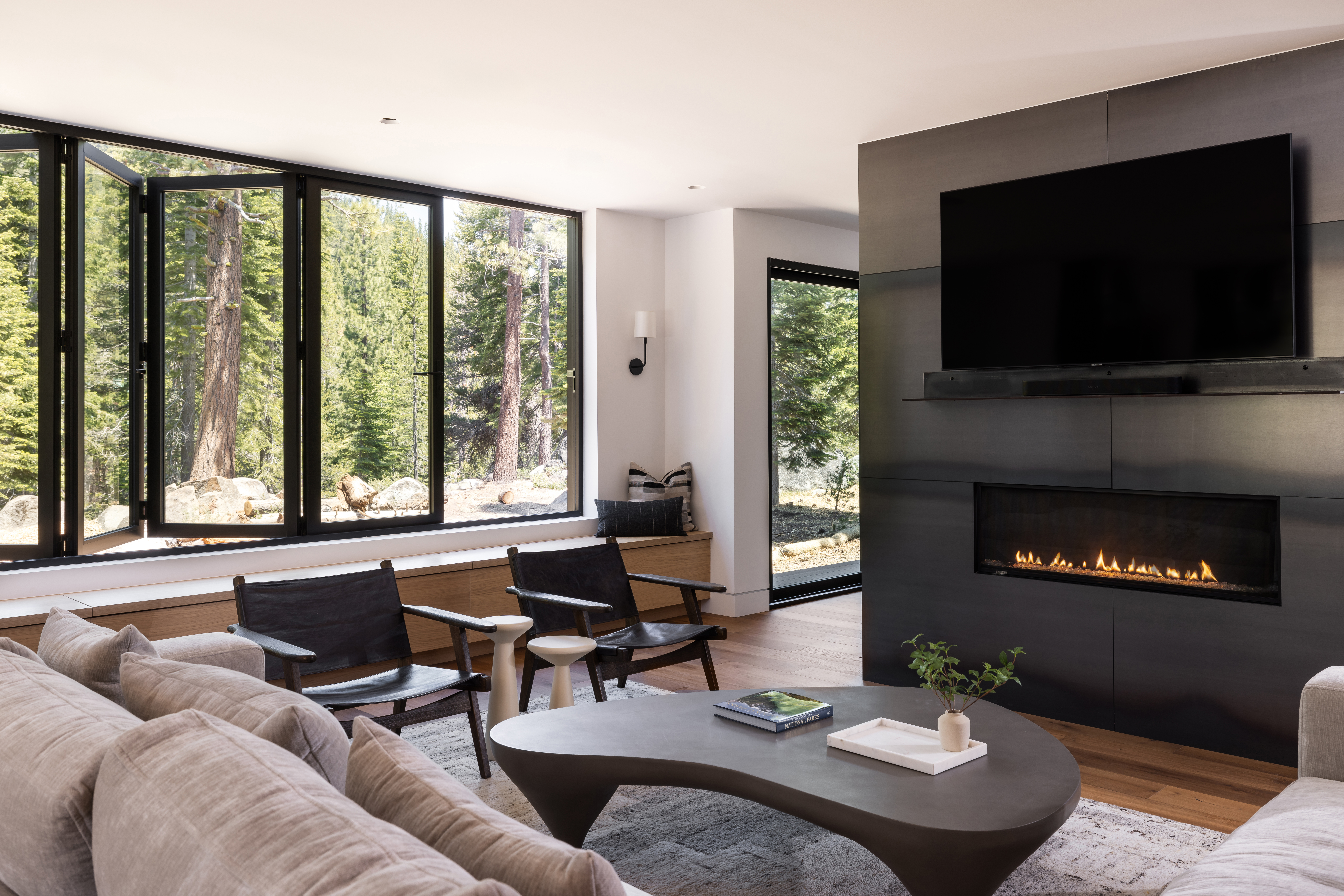 A contemporary living room of an Olympic Valley second home featuring a fireplace and expansive windows, offering ample natural light and stunning views.