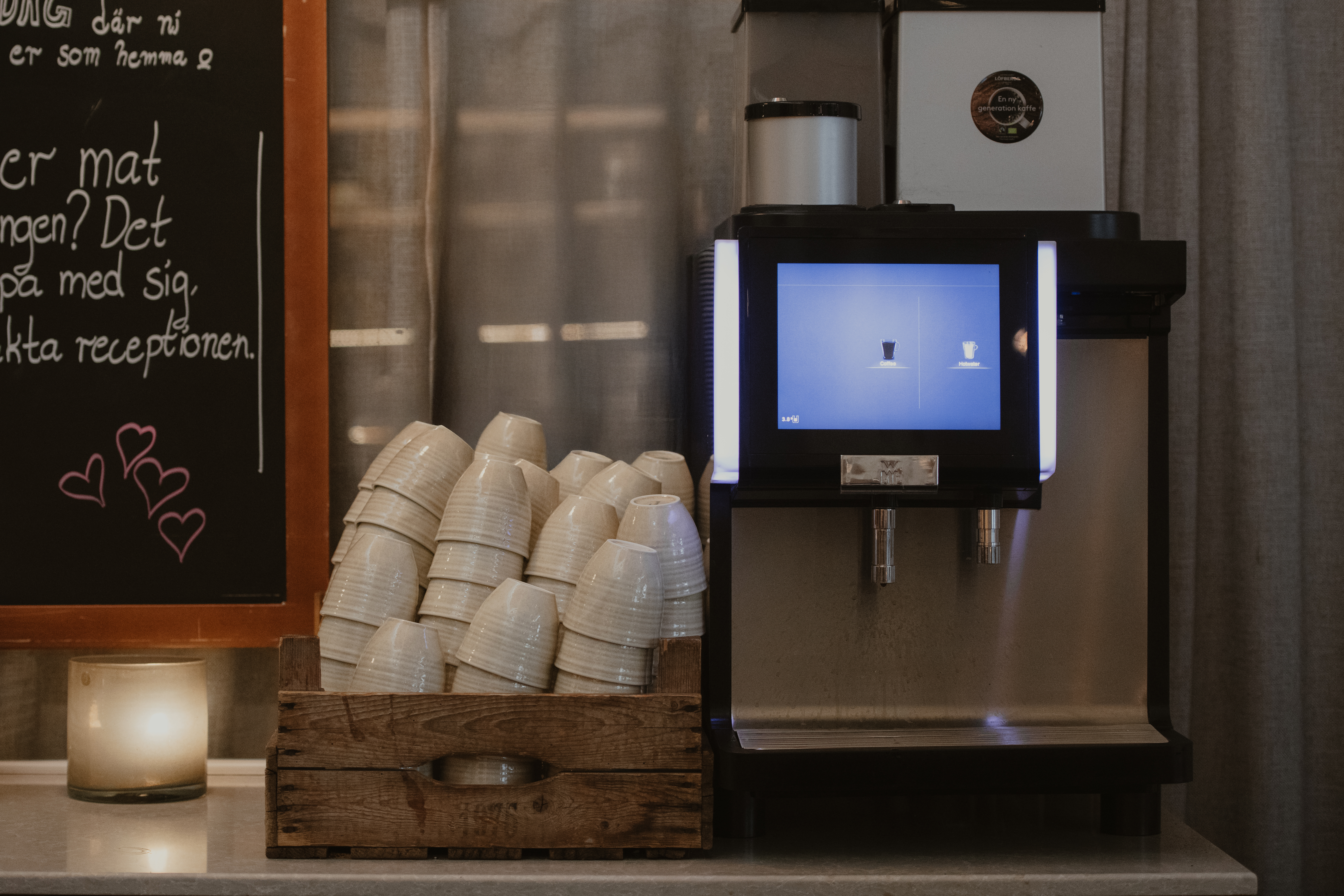 En kaffemaskin visar en meny bredvid en låda med staplade koppar på en bänk. I närheten står en griffeltavla med texten: "Har du mer mat i magen? Det kan du med sig fråga receperionen."