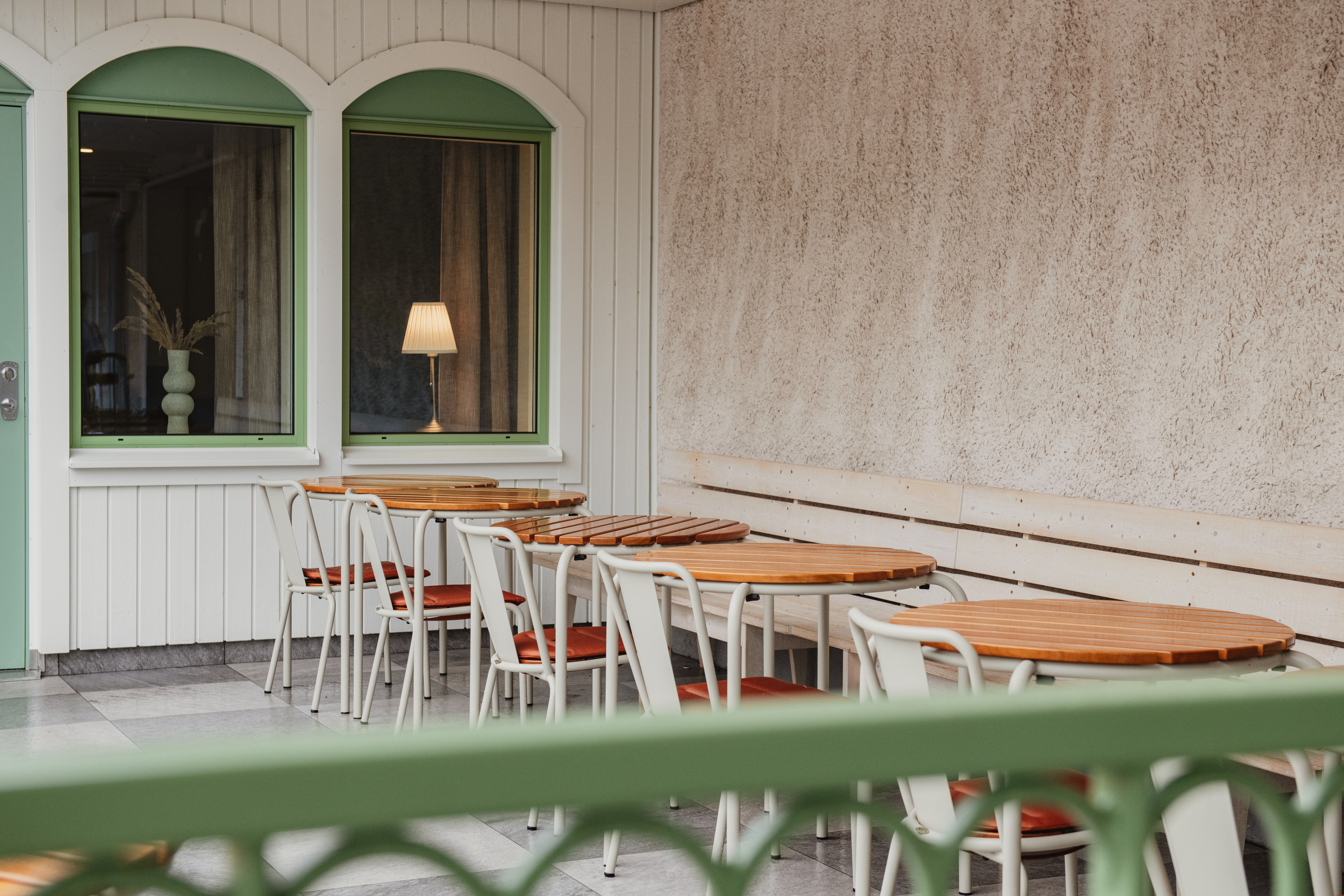 En indbydende udendørs terrasse med runde træborde og hvide stole, foran en bygning med grønne vinduesrammer.