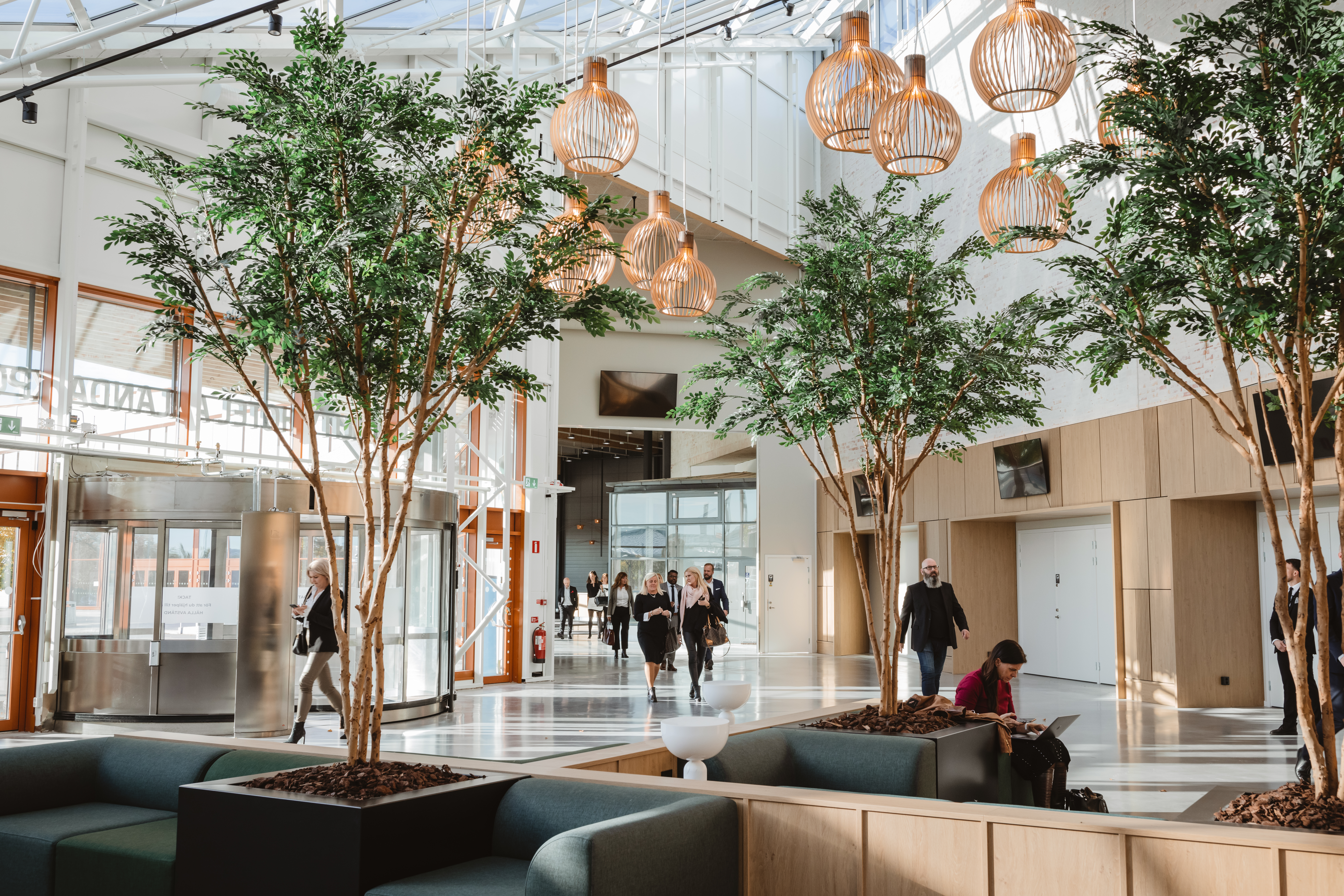 A bright, modern hotel lobby with people moving and relaxing. Indoor trees and unique lighting create a welcoming atmosphere. Arlanda.