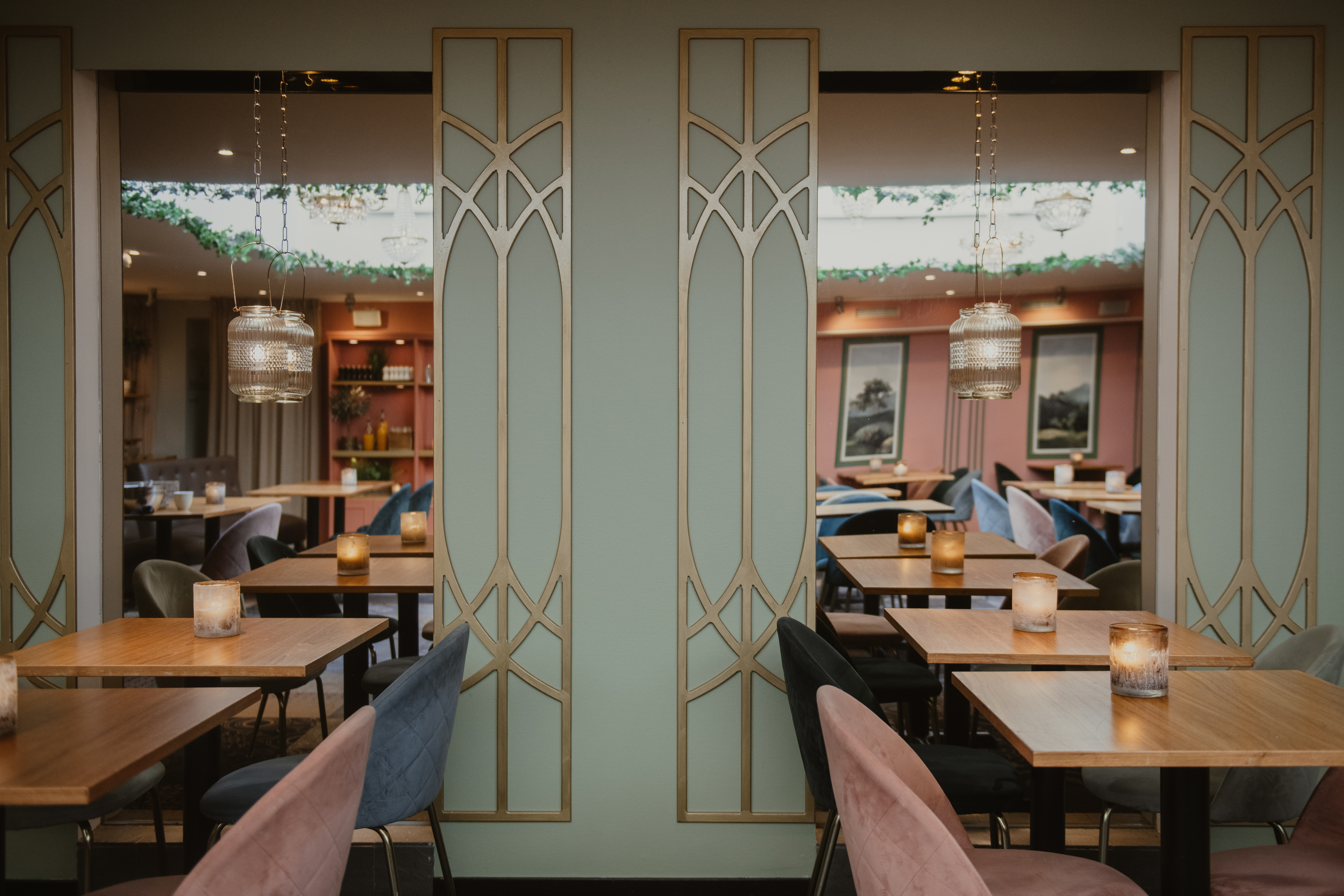 Bord med rosa och blå stolar är dukade med ljus i en stilren, mjukt belyst restaurang. Dekorativa avdelare med geometriska mönster separerar sittplatserna och förstärker den mysiga atmosfären.