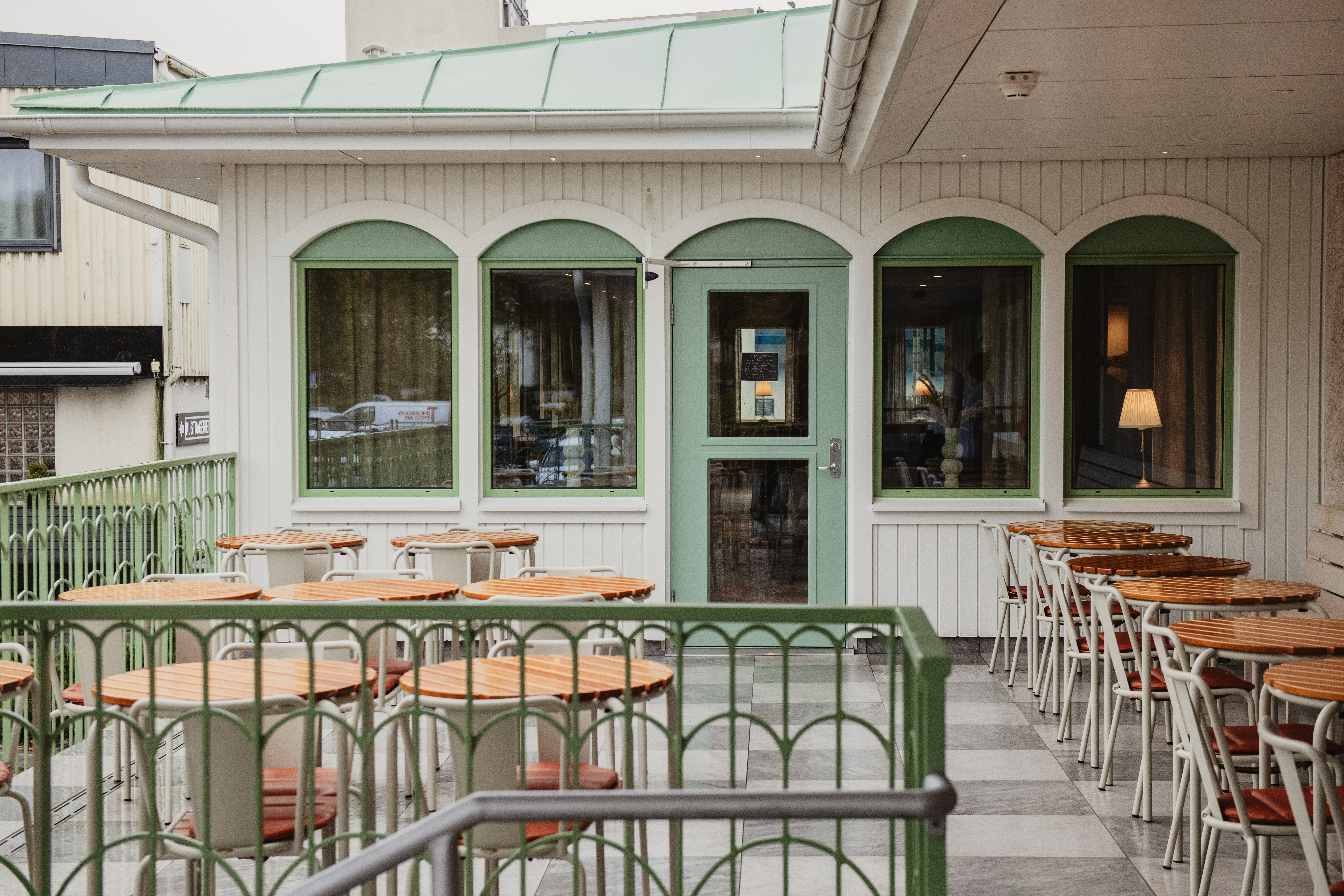 En indbydende udendørs caféterrasse med mange borde og stole, ved siden af en bygning med grønrammede vinduer og en dør.