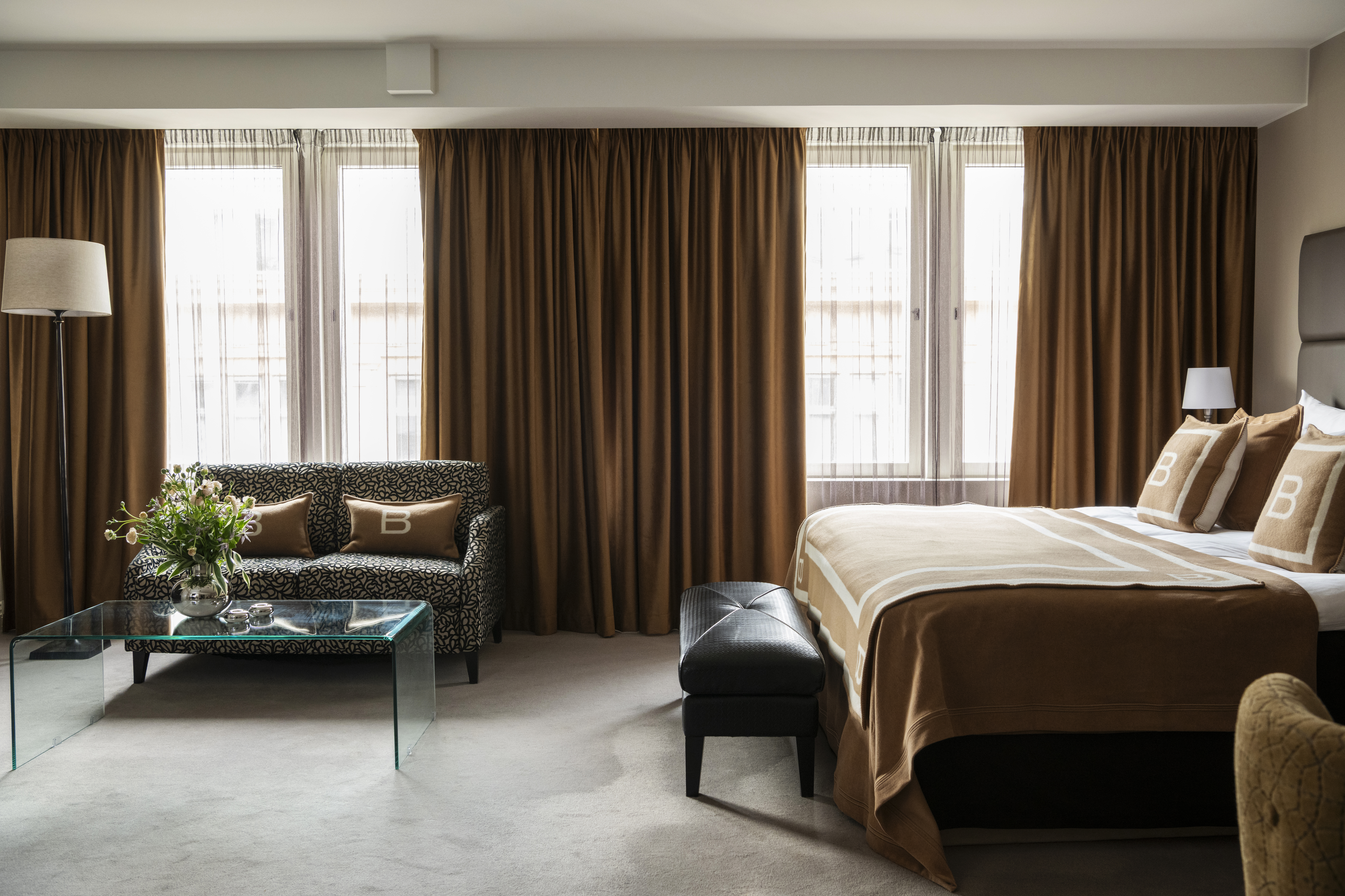 Bed with brown bedding is neatly made; large windows with closed brown curtains provide natural light. A patterned sofa and glass coffee table decorate the room.